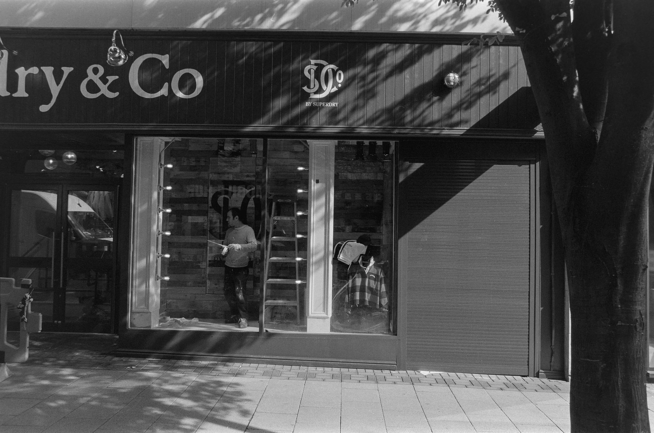 A storefront with a sign that reads 'ery & Co' and a smaller logo beneath it. Two men are visible through the glass window inside the store, one appears to be reading a newspaper. The exterior includes a large tree casting shadows on the building and