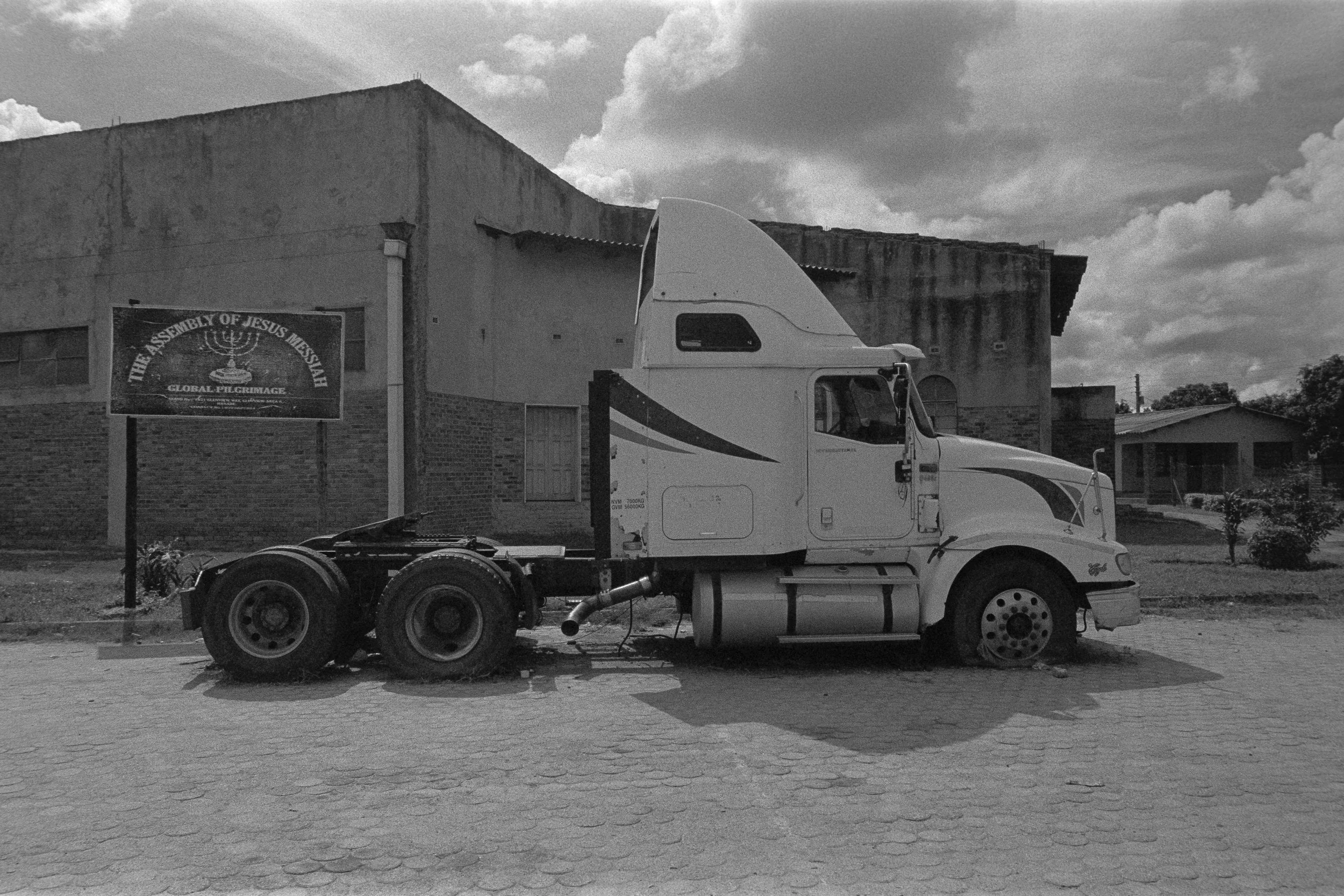 A black and white photo of an abandoned semi-truck without its trailer parked on a street in front of brick and concrete buildings. There is a sign on the building that reads 'The Assembly of Jesus Mexsih' and 'Global Revival.