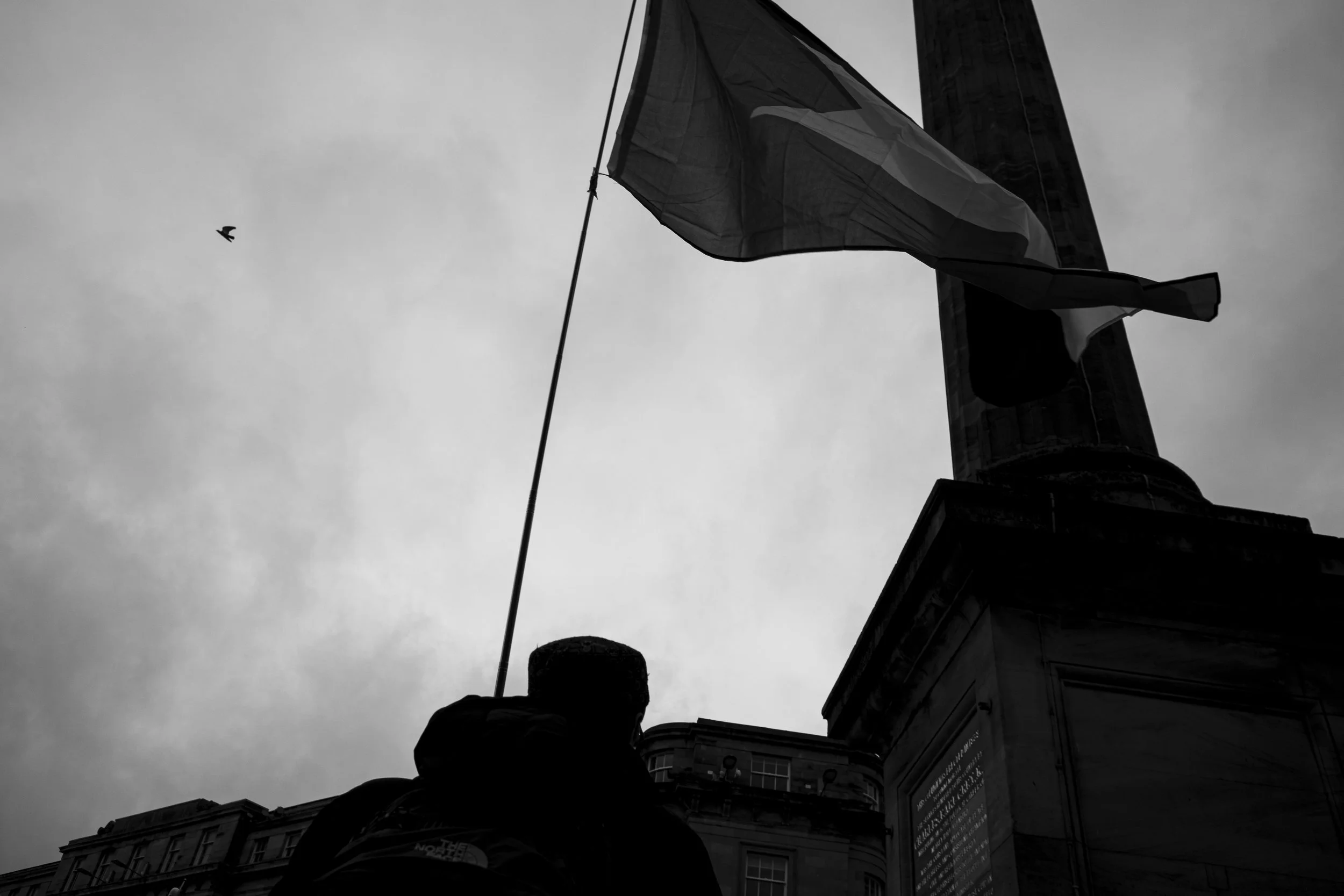 Black and white photo of a flag on a tall flagpole, with cloudy sky in the background. A person is seen silhouette in front of the flag, with part of a building also visible.