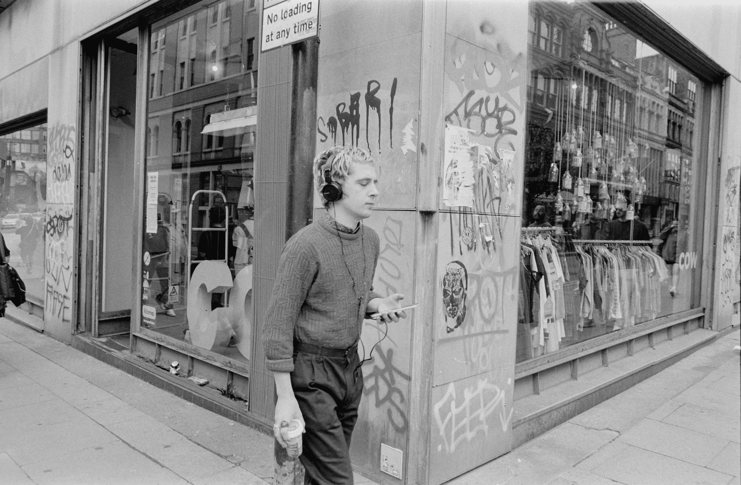 A young man with light-colored hair, wearing a sweater and dark pants, is walking past a graffiti-covered storefront while listening to music with headphones and holding a phone in one hand and a water bottle in the other, on a city sidewalk.