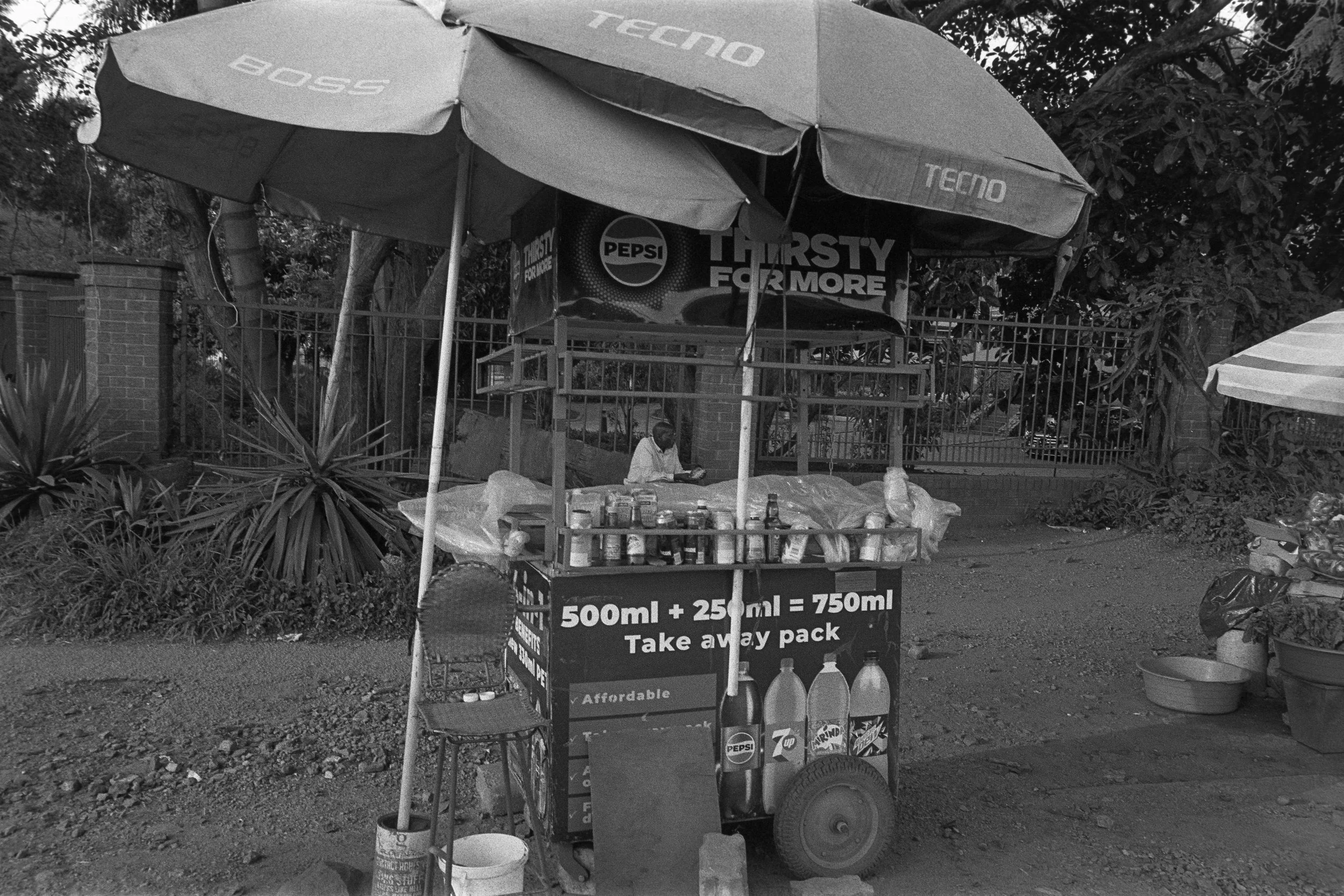 A street vendor stand with a large umbrella selling bottled drinks, with a sign indicating a take-away pack of 750ml.