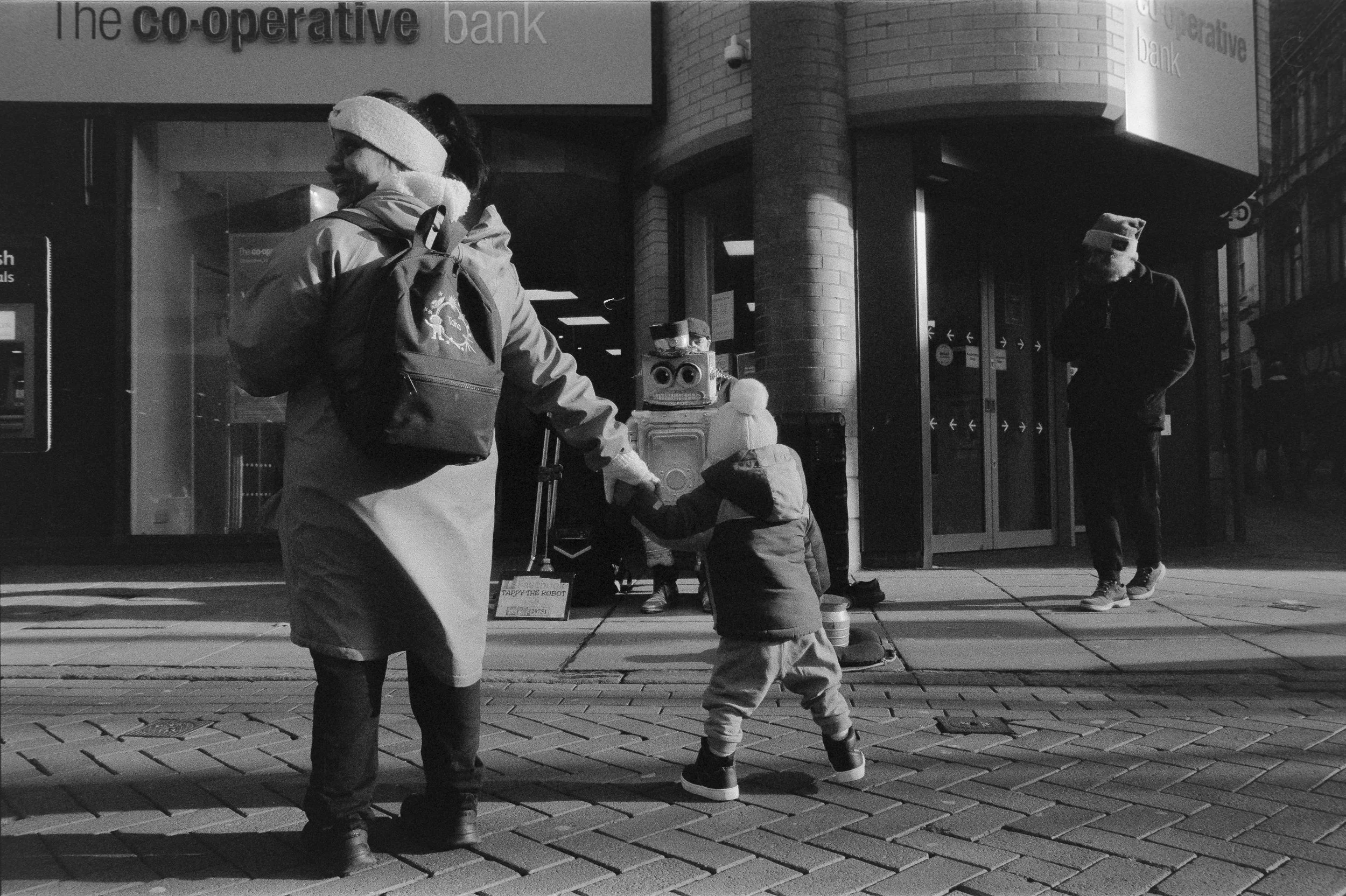 A woman and a child reaching out to a robot on the sidewalk outside a bank, with a man in the background talking on his phone.