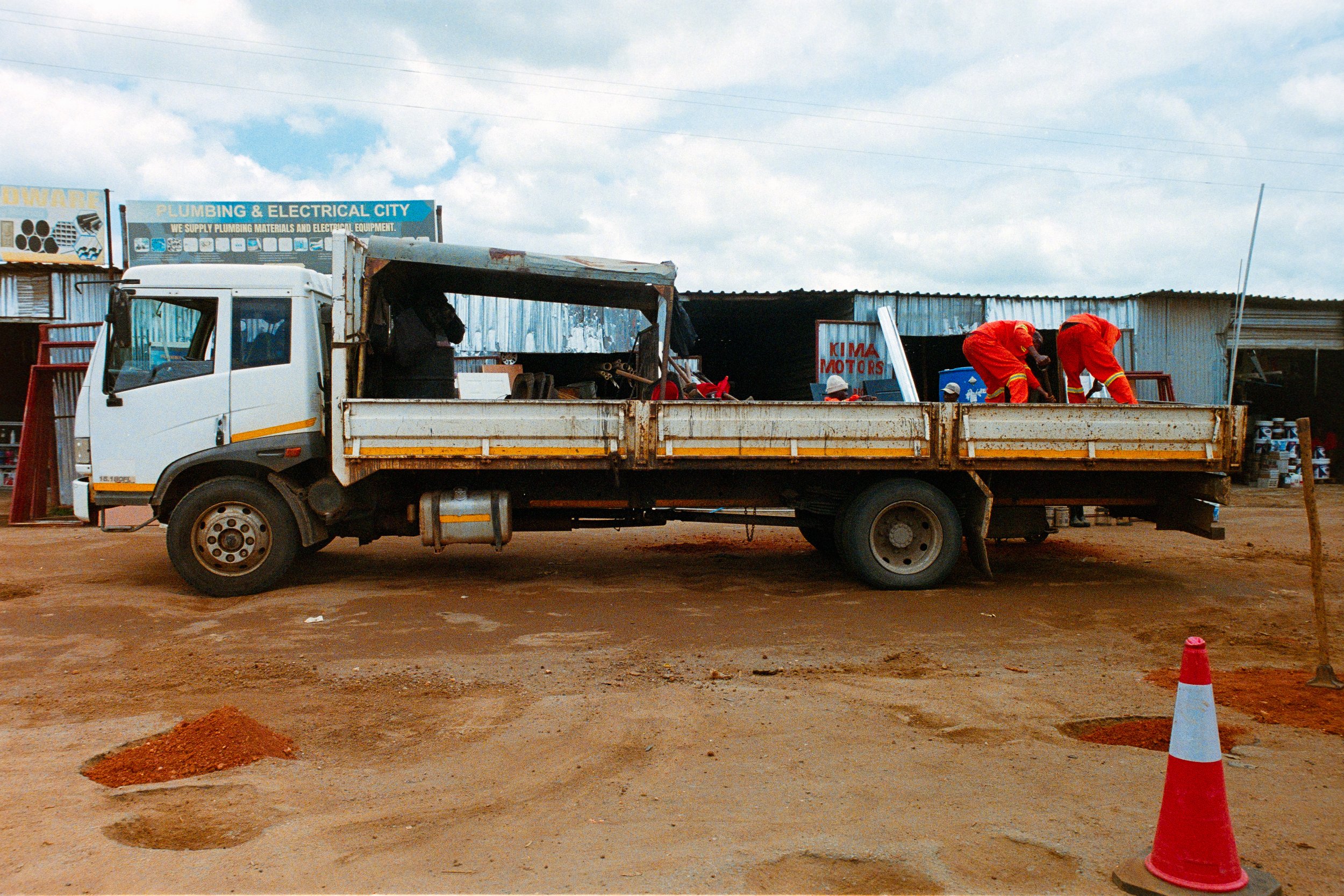 A white flatbed truck parked on a dirt road with two workers in orange uniforms working on the back. The background shows a building with signs, including one that says 'Plumbing & Electrical City' and another that says 'Kima Motors.' There is a traf