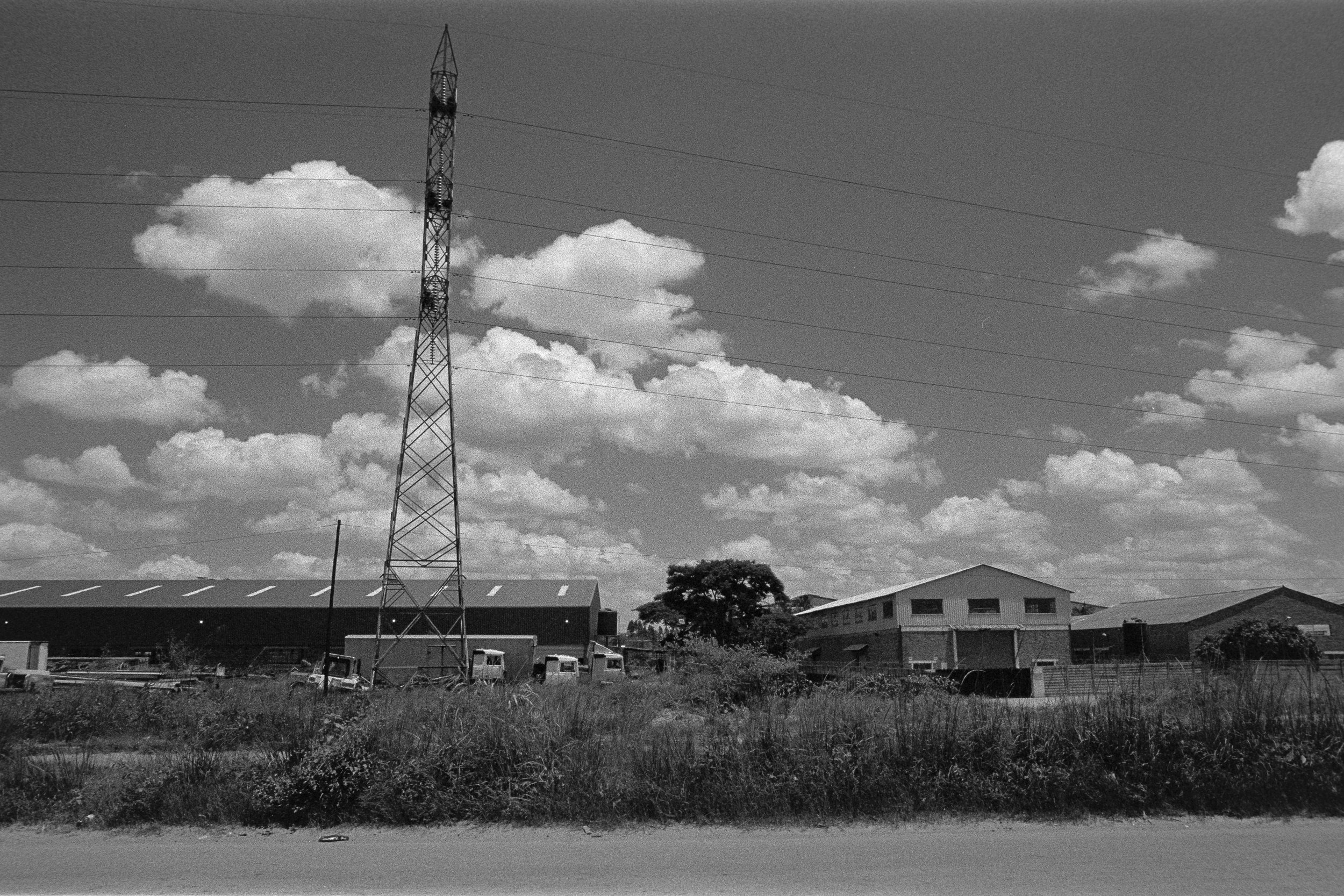 Black and white photo of a rural industrial area with a tall electric transmission tower, warehouses, and some vehicles, under a partly cloudy sky.