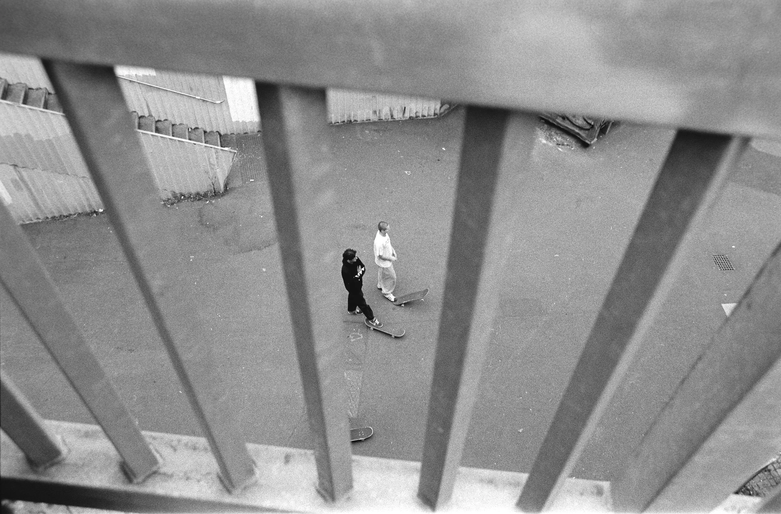 Black and white photo taken from above shows two skateboarders standing on a paved surface, viewed through metal railings. One skateboarder wears a dark hoodie and pants, the other a light top and sweatpants. The scene includes a small-gated area and