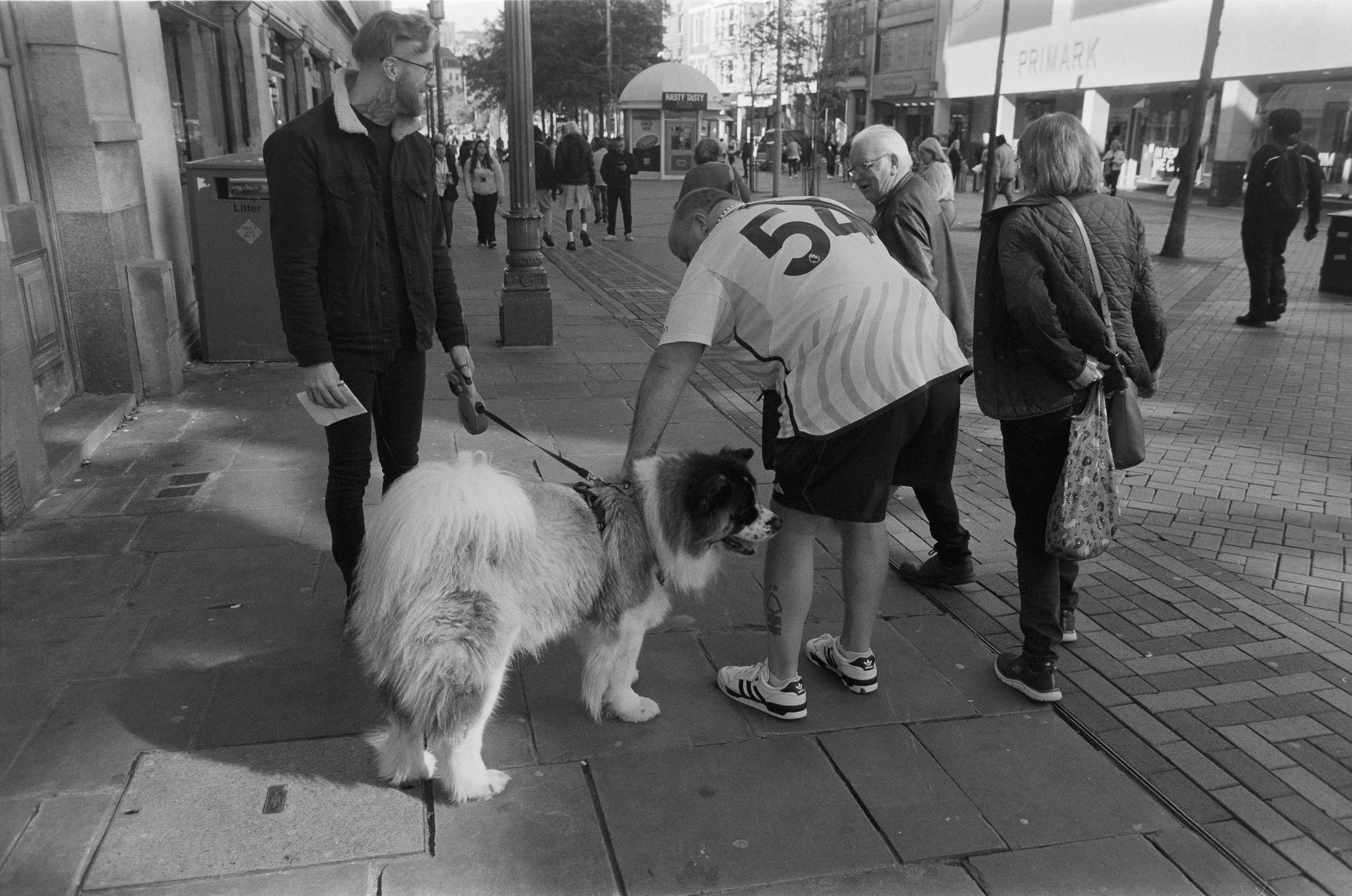 A group of people on a city sidewalk, with a man petting a large fluffy dog, and another man holding its leash. Other pedestrians are walking in the background, along with storefronts and a bus stop.