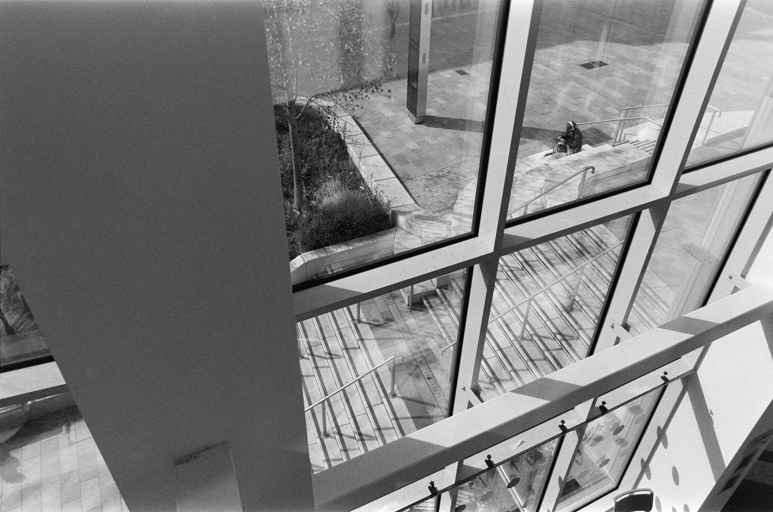 Black and white photo of a modern building's interior with large glass windows showing stairs outside. A person is sitting on stairs outside, viewed from above.