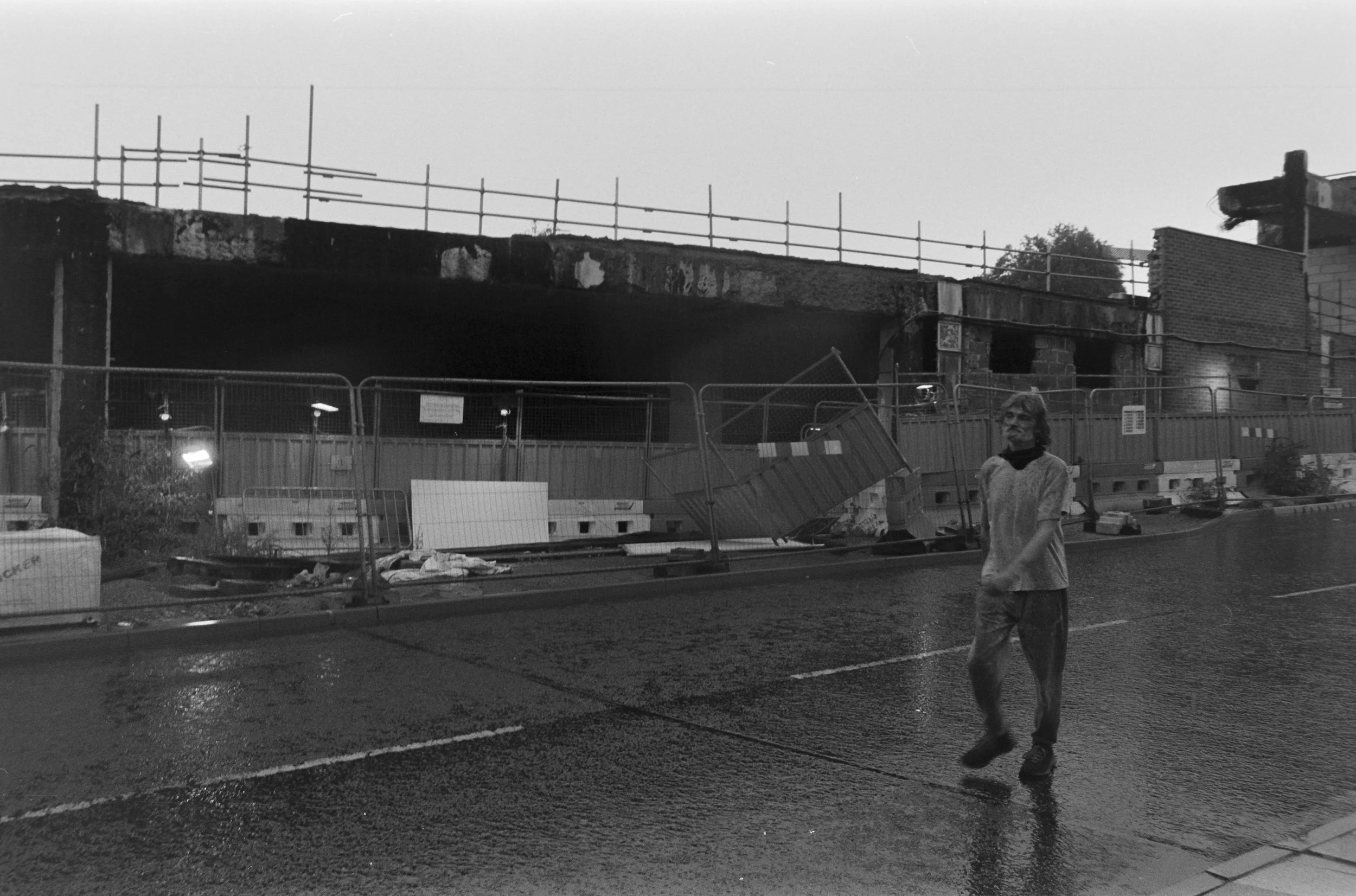 A person walking on a wet street near a construction site with a toppled fence and a burned building in the background, black and white photo.