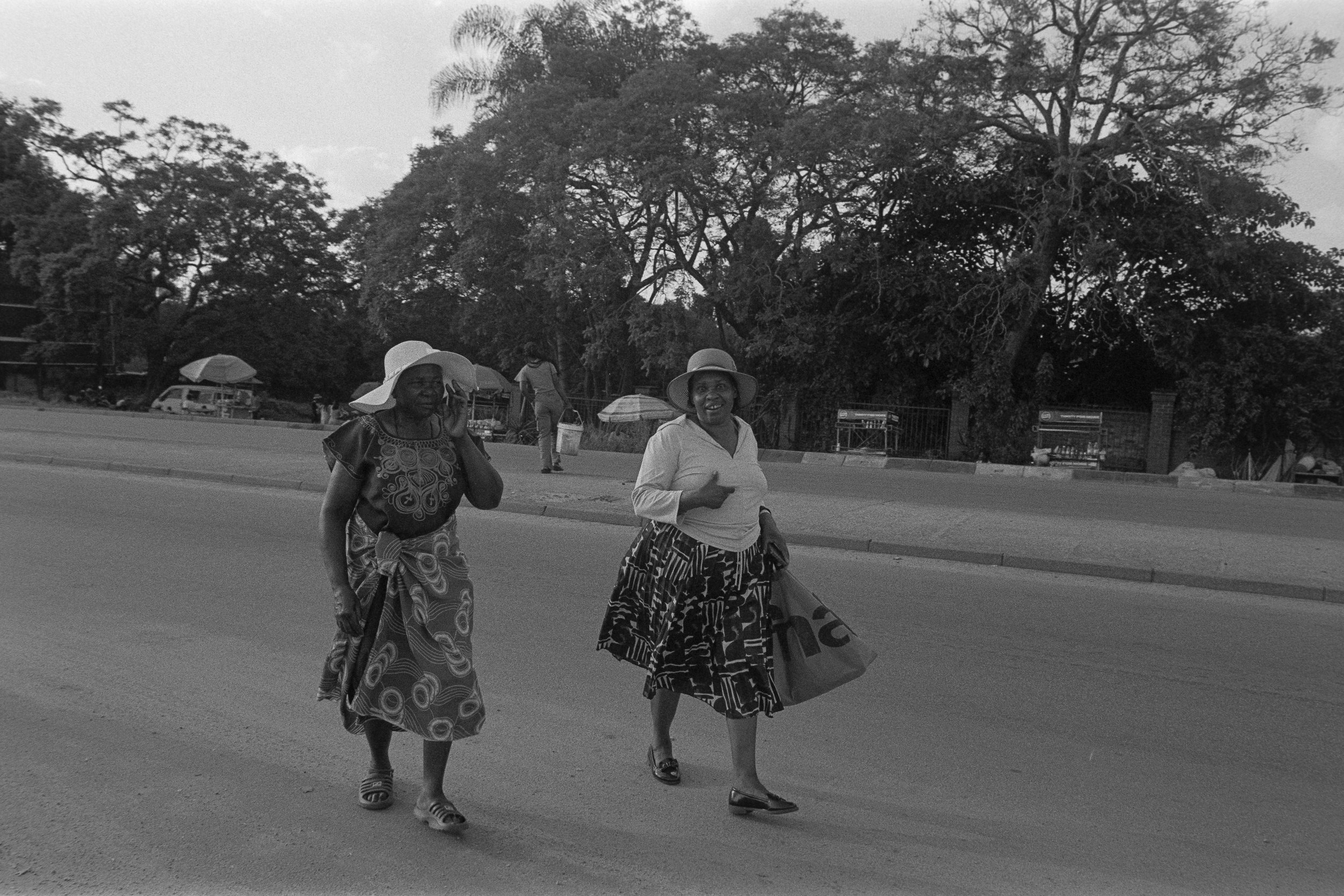 Two women wearing skirts and hats walking on a street, with trees and a person carrying a bag in the background.