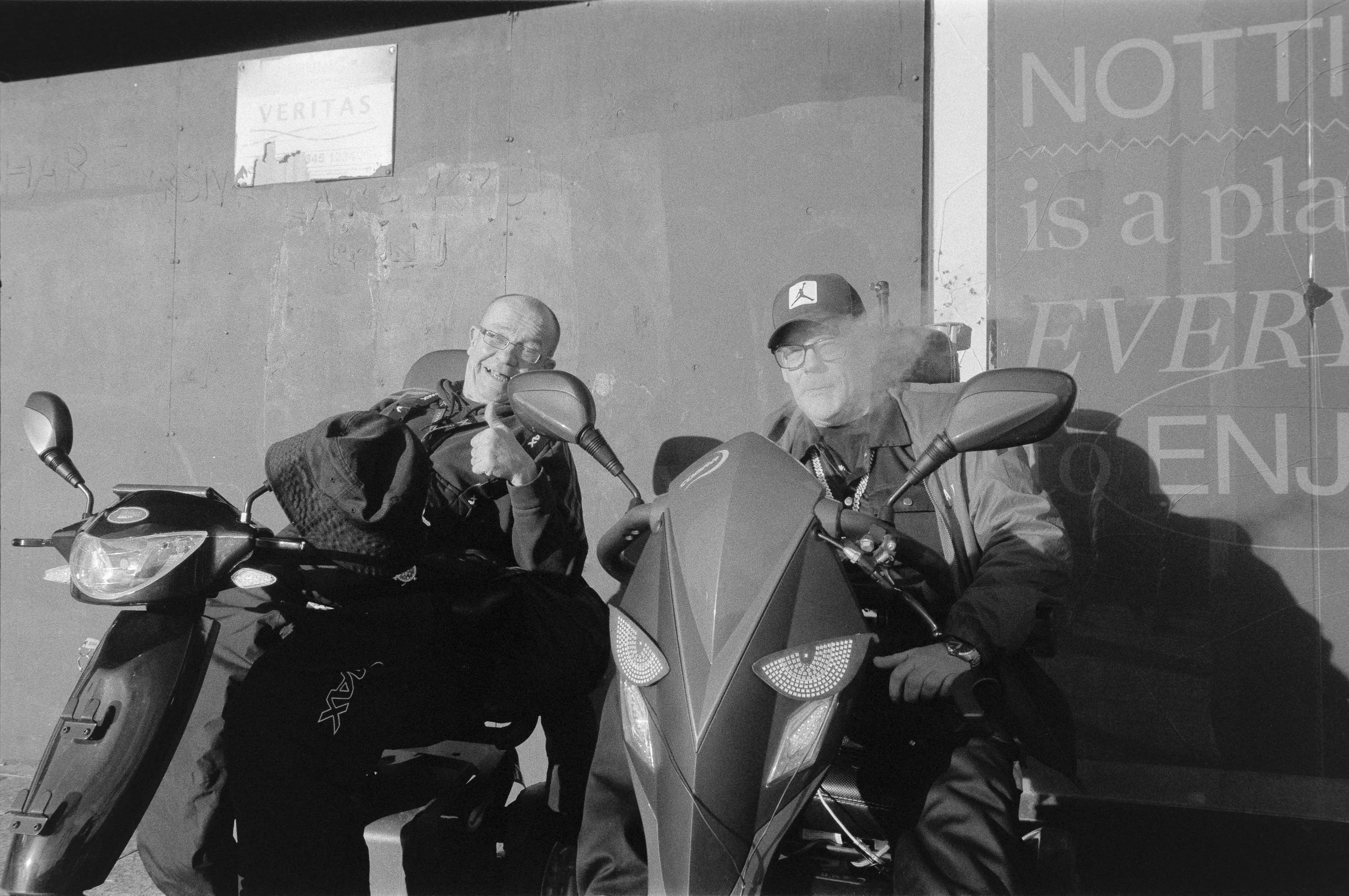 Two older men sitting on motorcycles outside a building, one giving a thumbs-up and smiling, the other smoking and looking down. The photo is black and white.