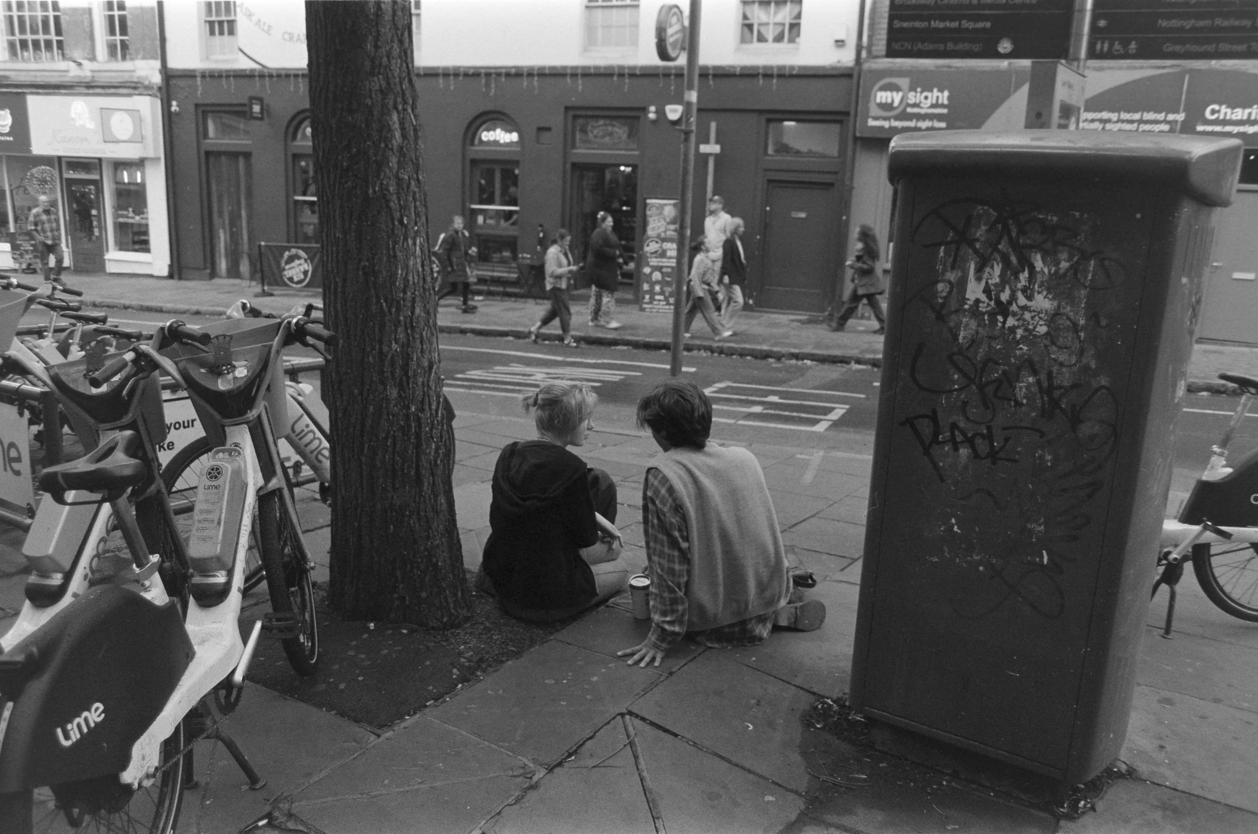 Two people sit on the sidewalk under a tree, facing each other, with bicycles nearby. Behind them, a street scene shows pedestrians walking on the sidewalk and across a crosswalk. There is a large, graffiti-covered utility box on the right and a buil