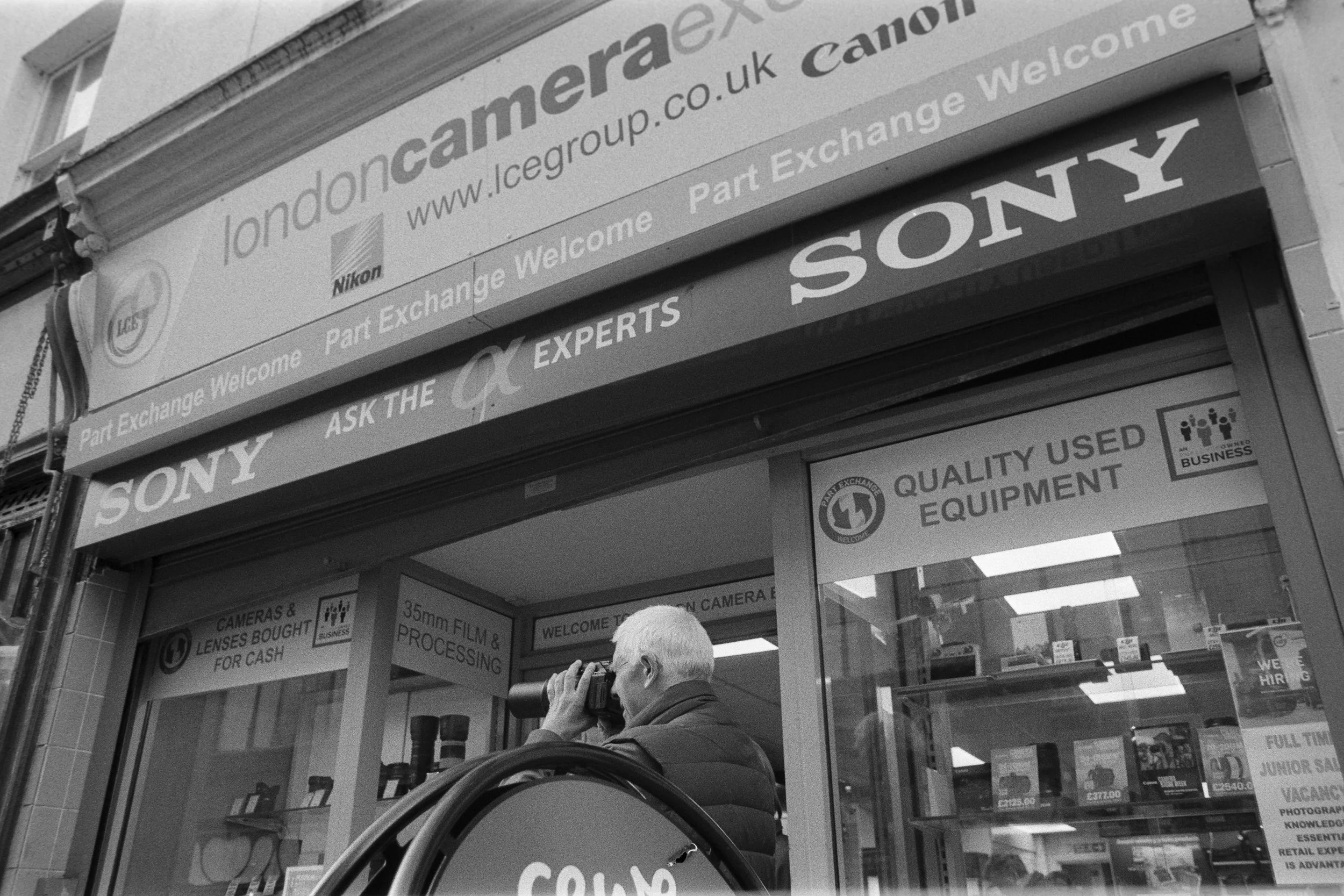 A man looking through binoculars outside a camera shop in London, with signs advertising used equipment, camera film processing, and camera lens sales. The shop has signs for brands like Sony and Canon.