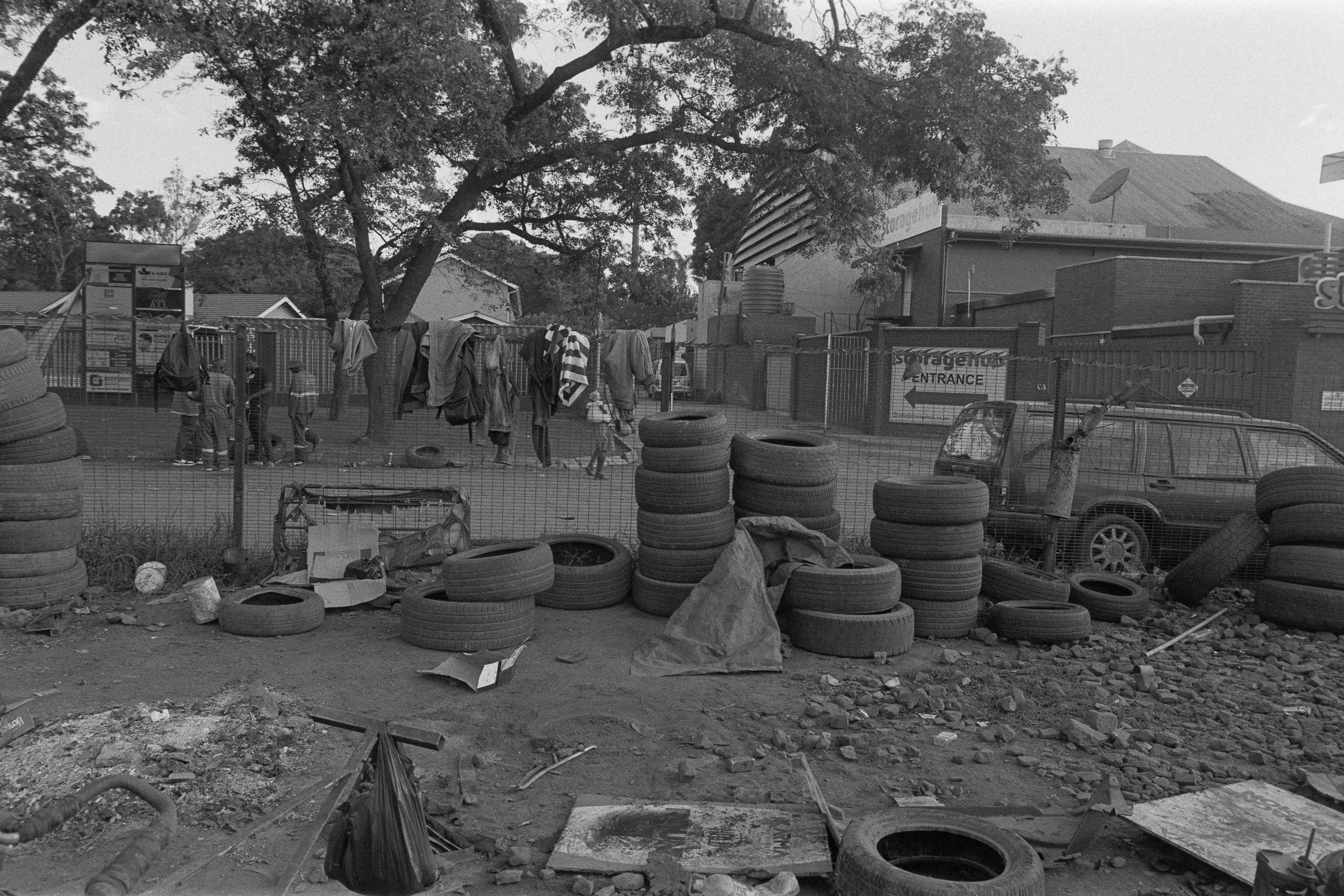 A black and white photo of a neglected outdoor area with scattered tires, discarded boxes, and debris. In the background, people are standing behind a fence, near clothes hanging to dry and a tree with full foliage.
