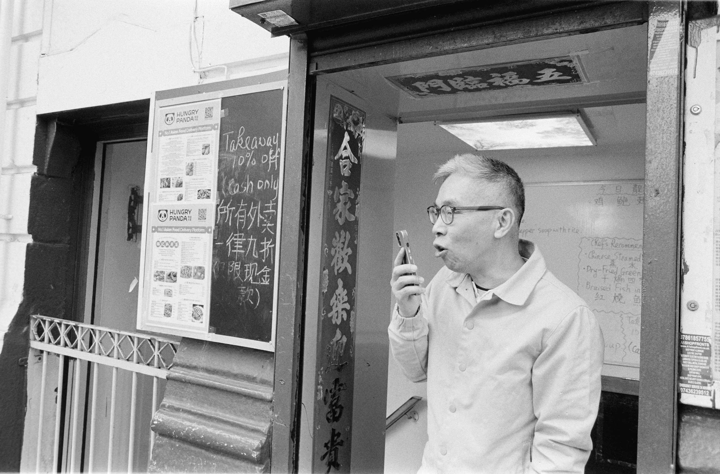 A man with glasses and a light-colored jacket standing outside a restaurant, speaking into a phone, with menus and signs on the wall behind him.