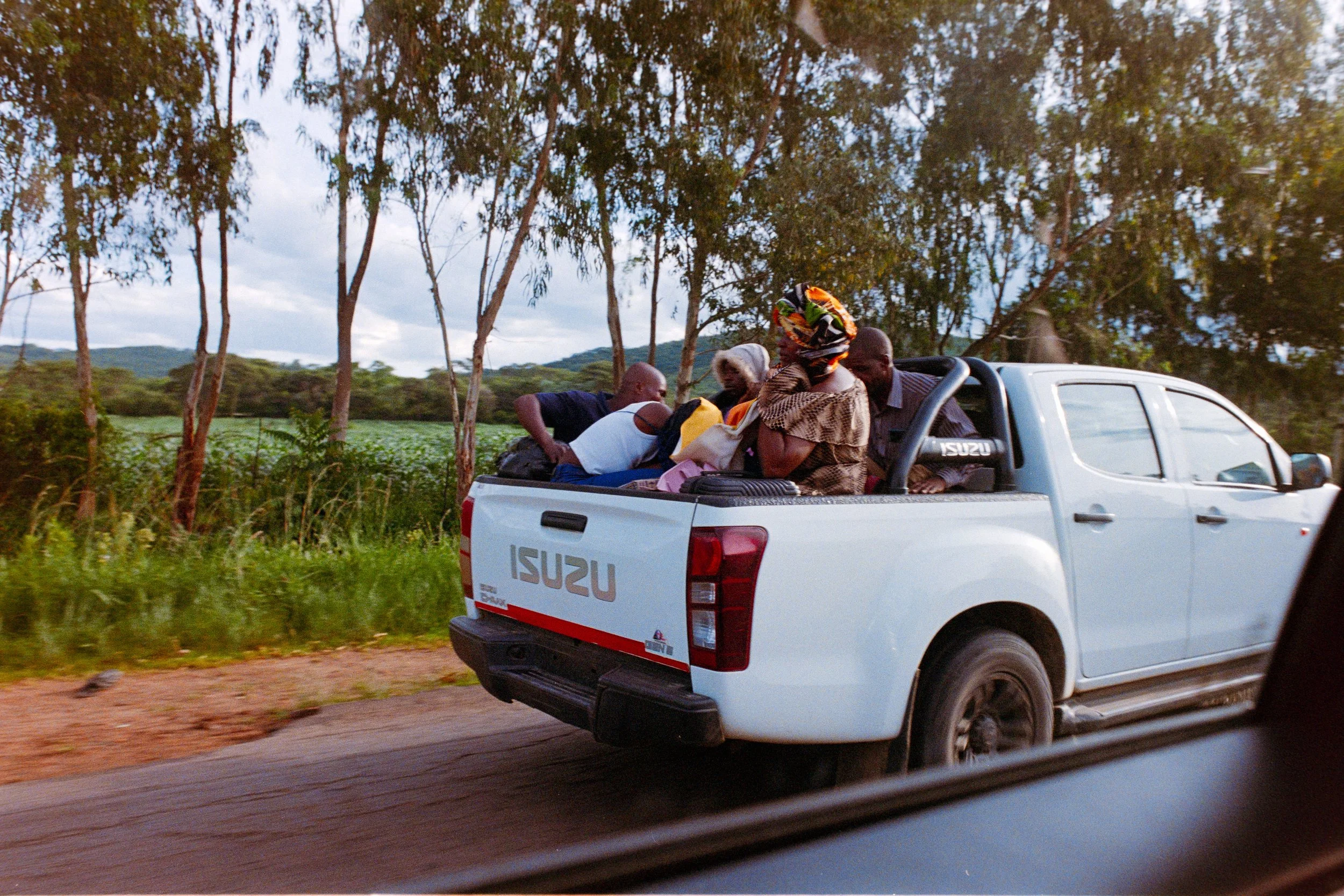 People riding in the back of a white Isuzu pickup truck on a rural road with trees and fields in the background.