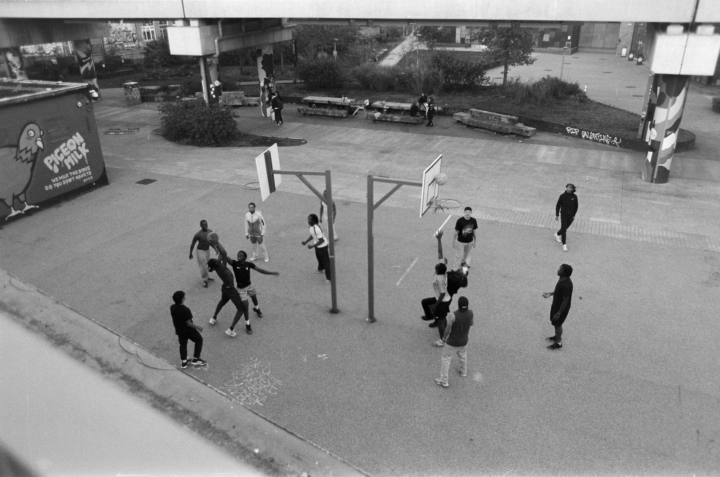 Children playing basketball on an outdoor court with some adults nearby in an urban setting, graffiti on walls, benches and a painted pole, with a large advertisement on the left.