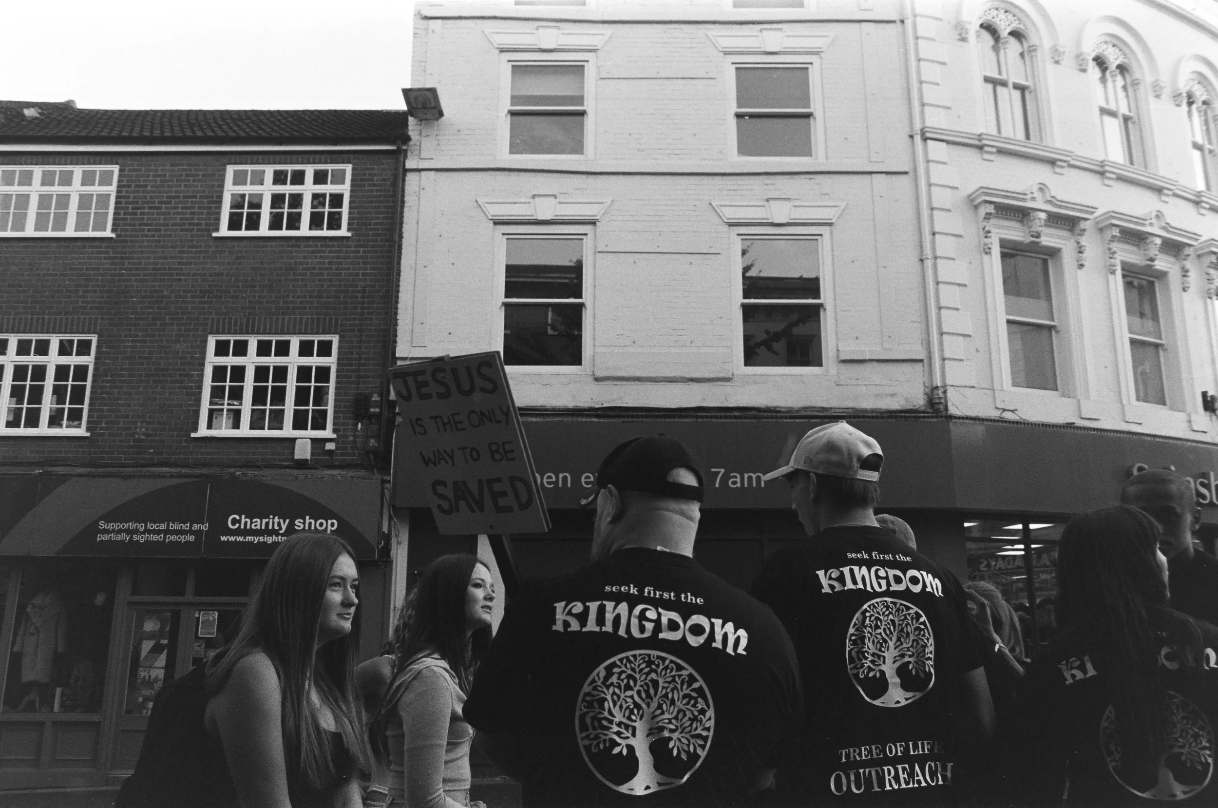 People participating in a street outreach event, with some holding a sign that reads 'Jesus is the only way to be saved.' They are standing in front of a row of buildings, including a charity shop and a church outreach group.
