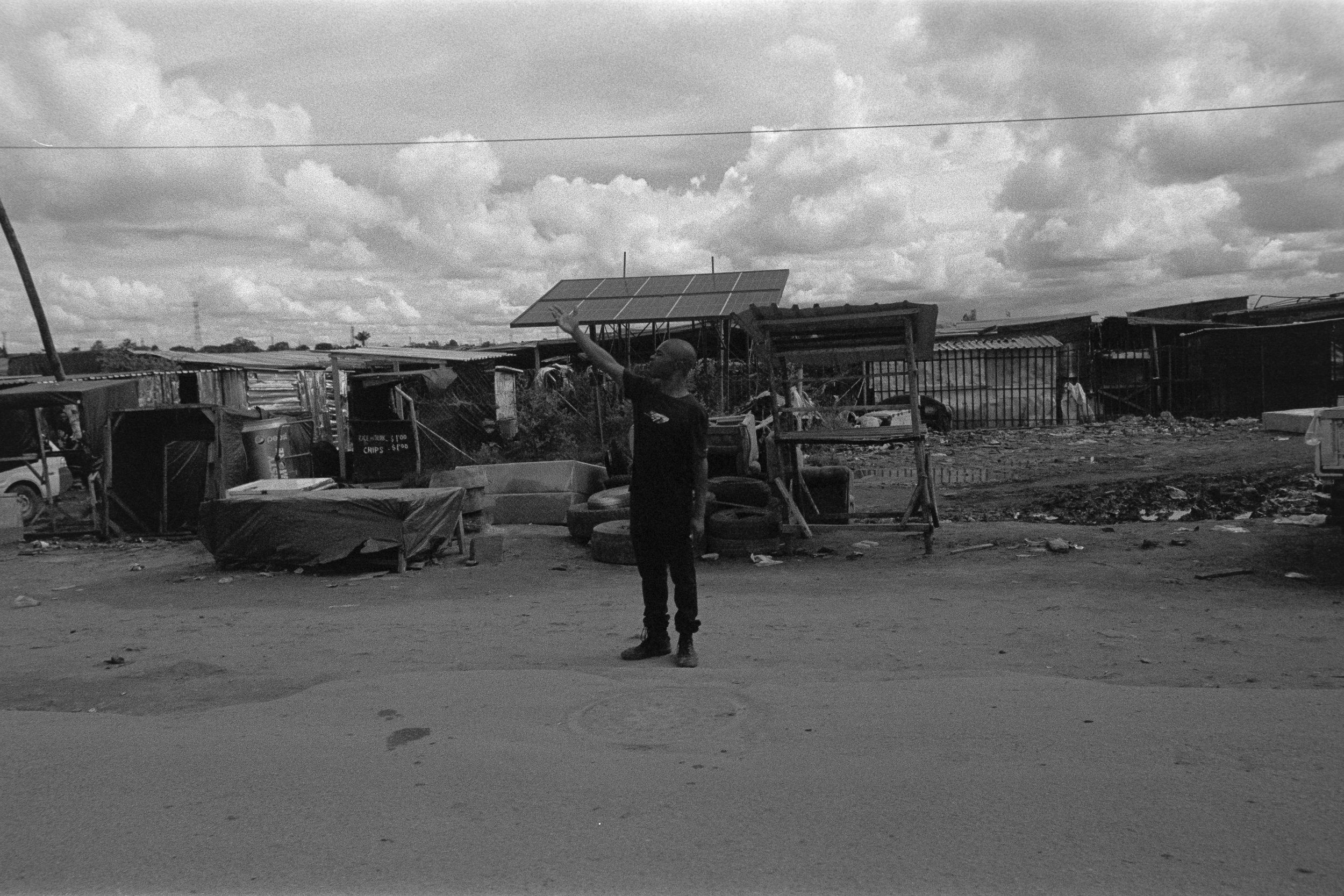 A man standing in a dirt road in a makeshift settlement with scrap materials, tires, and small structures under cloudy skies.