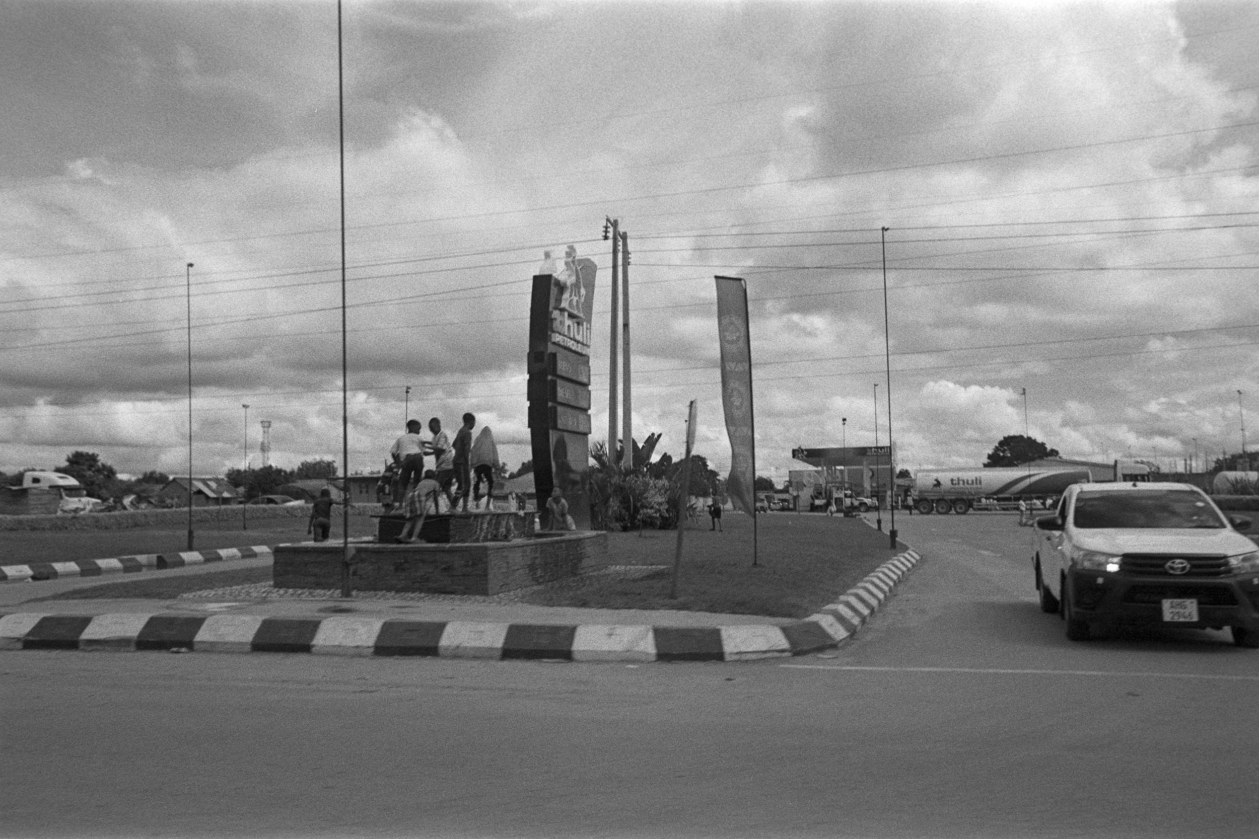 People standing and sitting on a roadside monument with signs and flags, surrounded by a curved road with vehicles including a pickup truck and a tanker, under a cloudy sky.