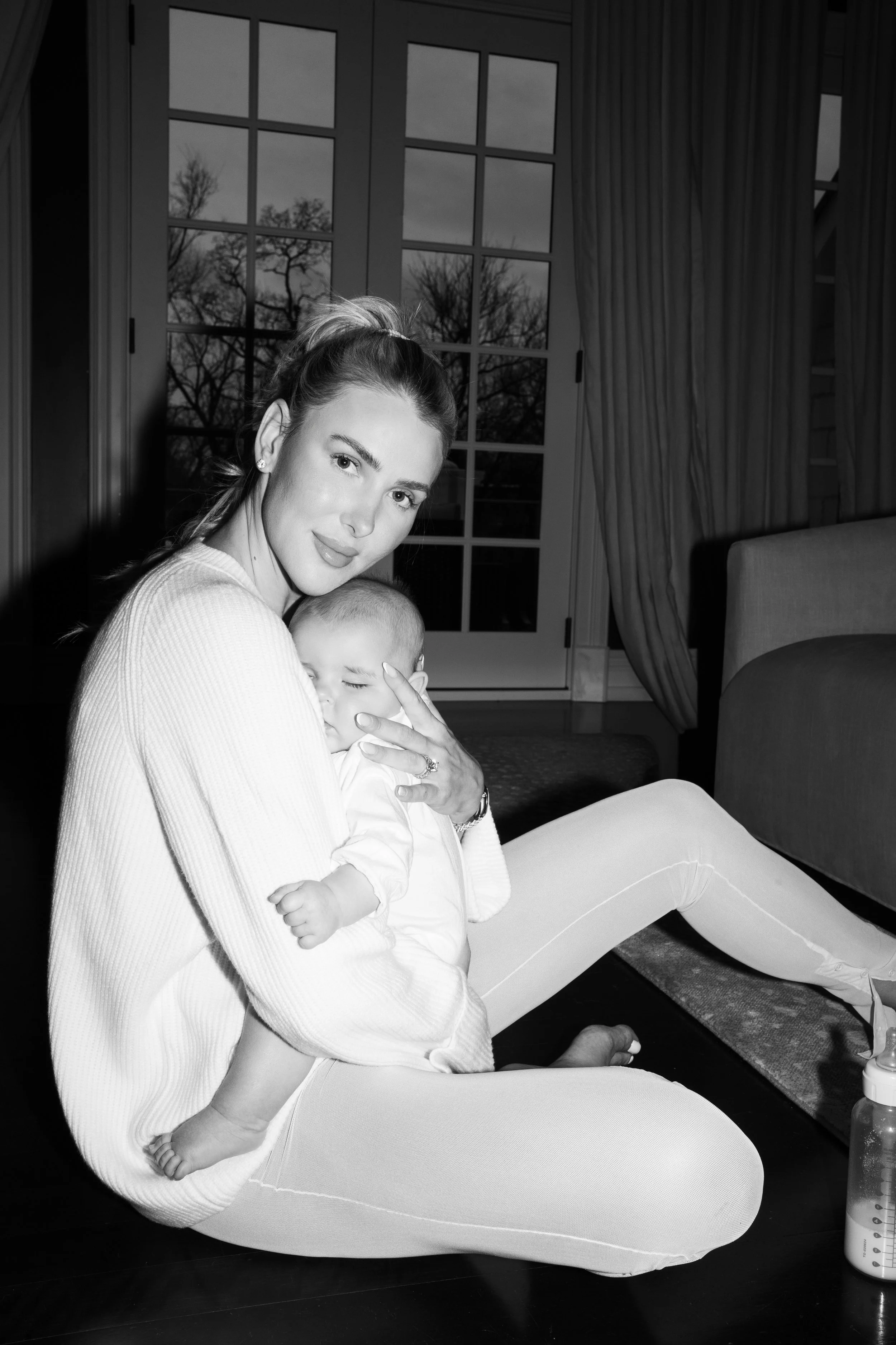 A woman sitting on the floor holding a sleeping baby against her chest, inside a room with large windows and curtains.