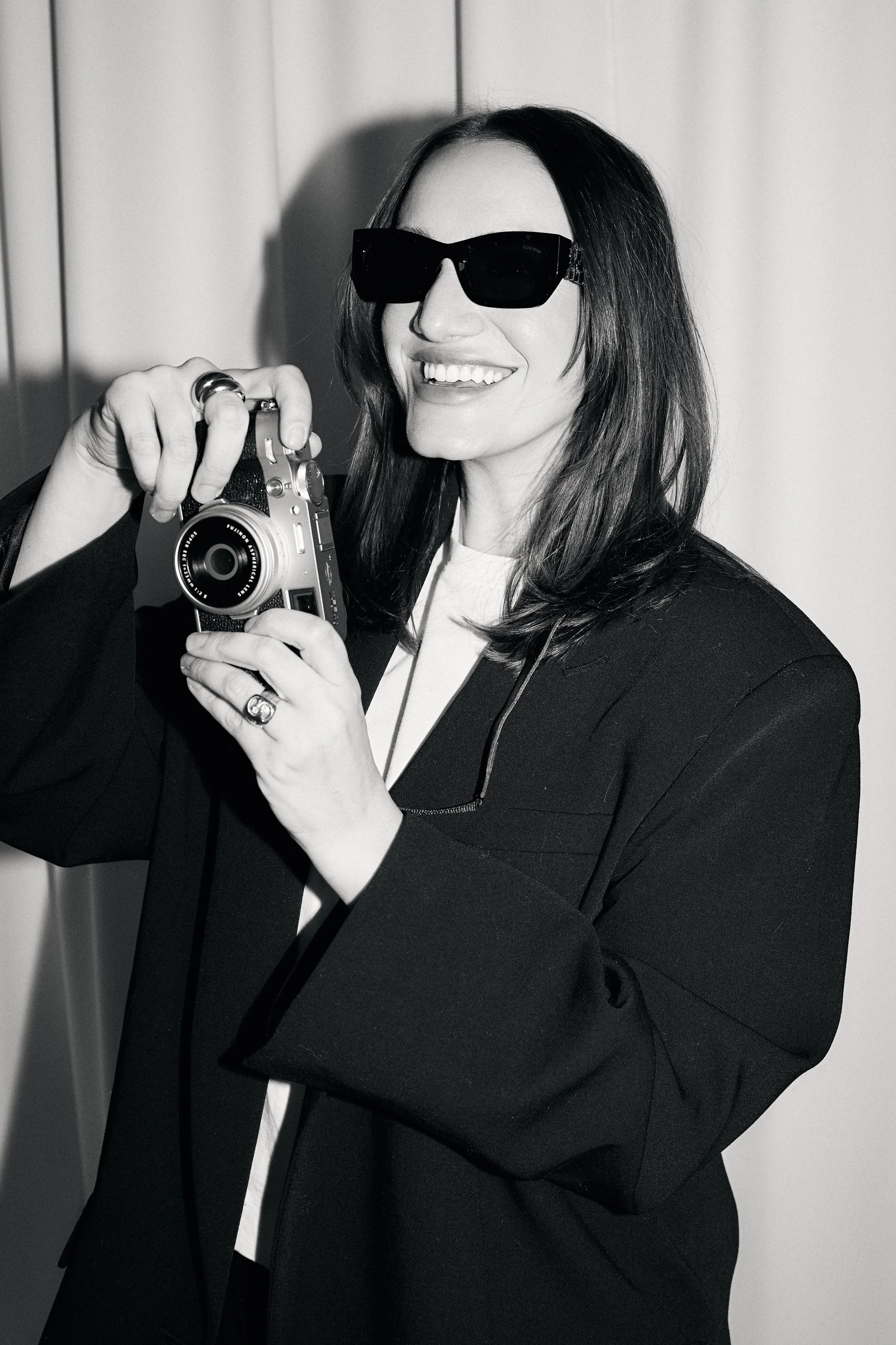 A woman smiling, wearing sunglasses and holding a camera, dressed in a dark blazer and white top.