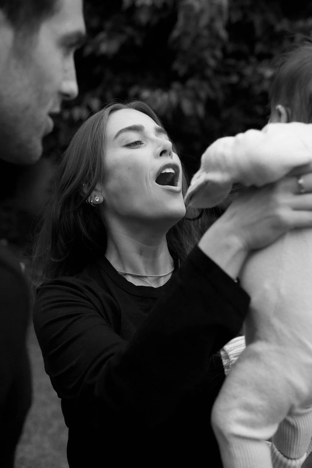 A woman with long hair and earrings holds a child in a track suit facing away. The woman appears to be speaking or singing towards the child, with her mouth open, and is in an outdoor setting with blurred foliage in the background.