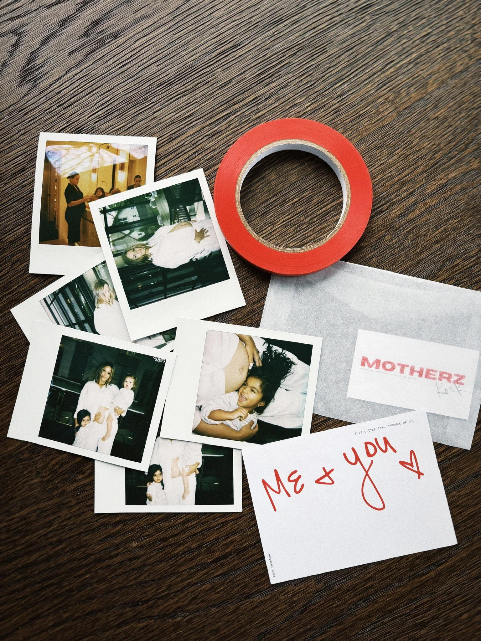 Collection of five Polaroid photos featuring women and children, a roll of red tape, and handwritten notes that say 'Me + you' with a heart, and a printed note labeled 'Motherz Rock' on a wooden table.