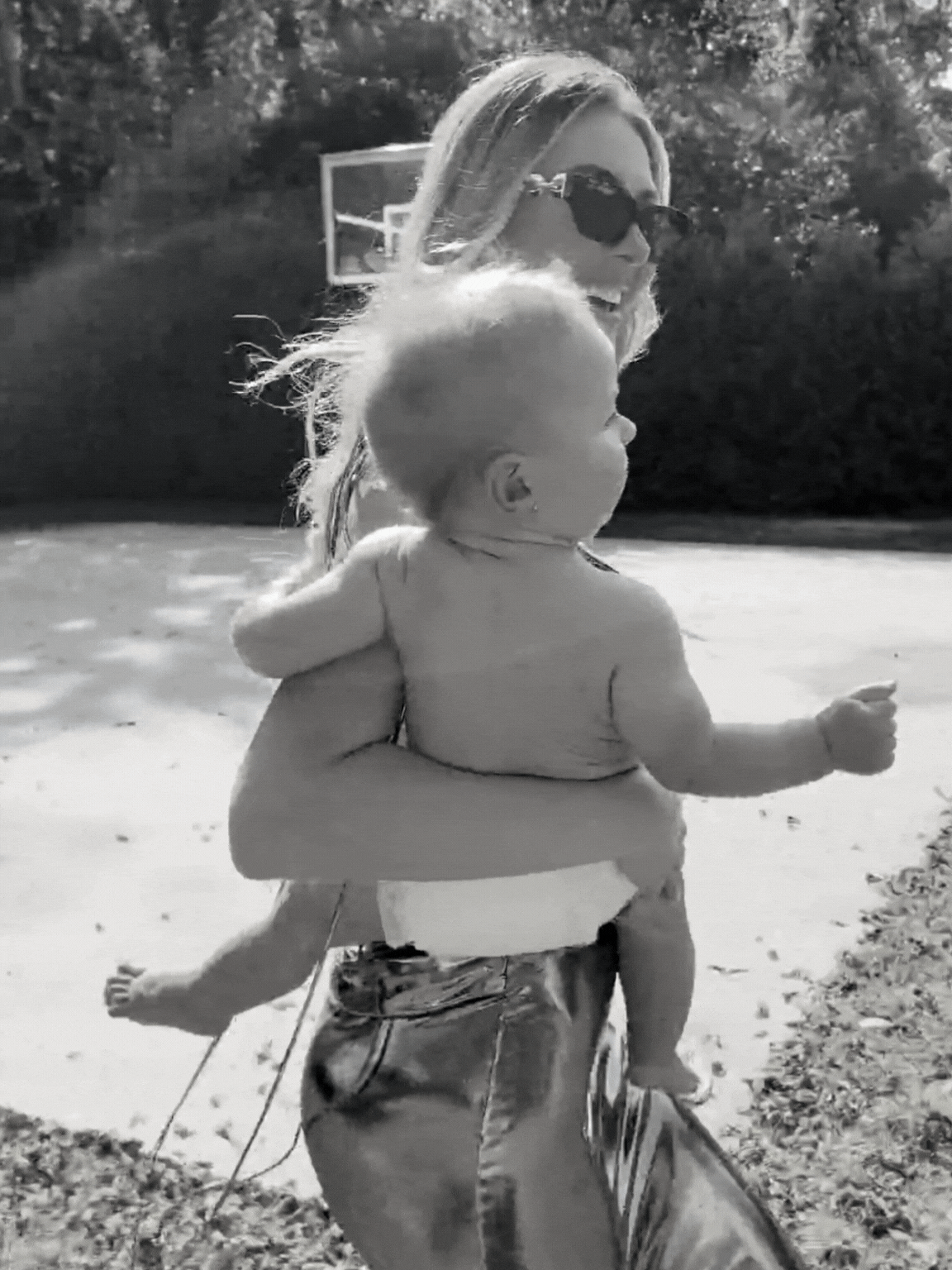 A woman wearing sunglasses is holding a young child, and they appear to be running outside on a sunny day, with a basketball hoop and trees in the background.