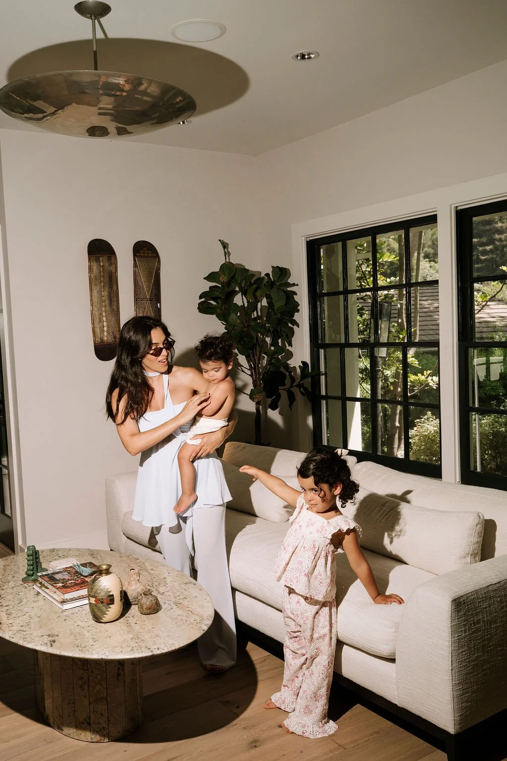 A woman holding a young boy in a white dress, playing with another young girl in pajamas in a living room with large windows, a white armchair, and a marble coffee table.