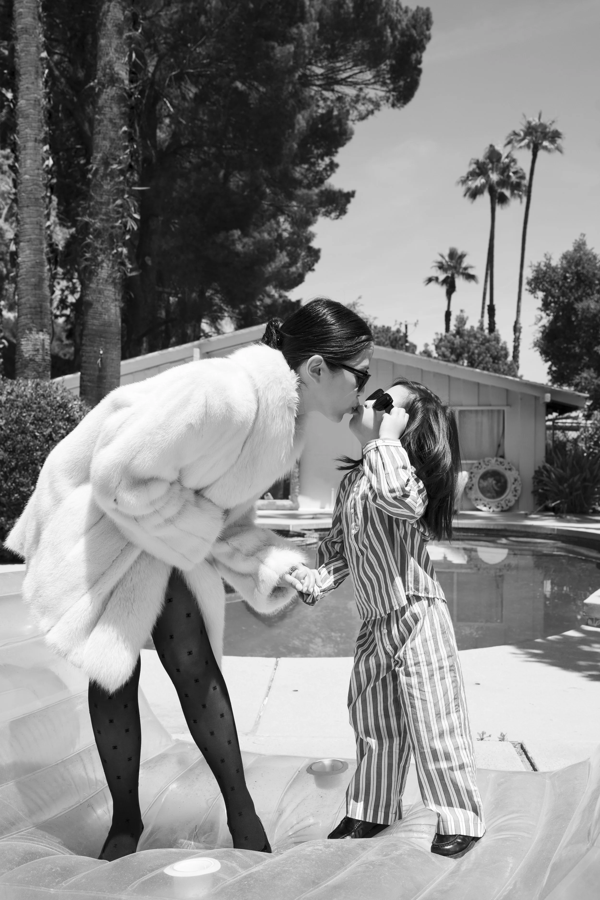 A woman and a young girl sharing a kiss outside near a pool, with trees and tall palm trees in the background, both dressed in fashionable clothing.