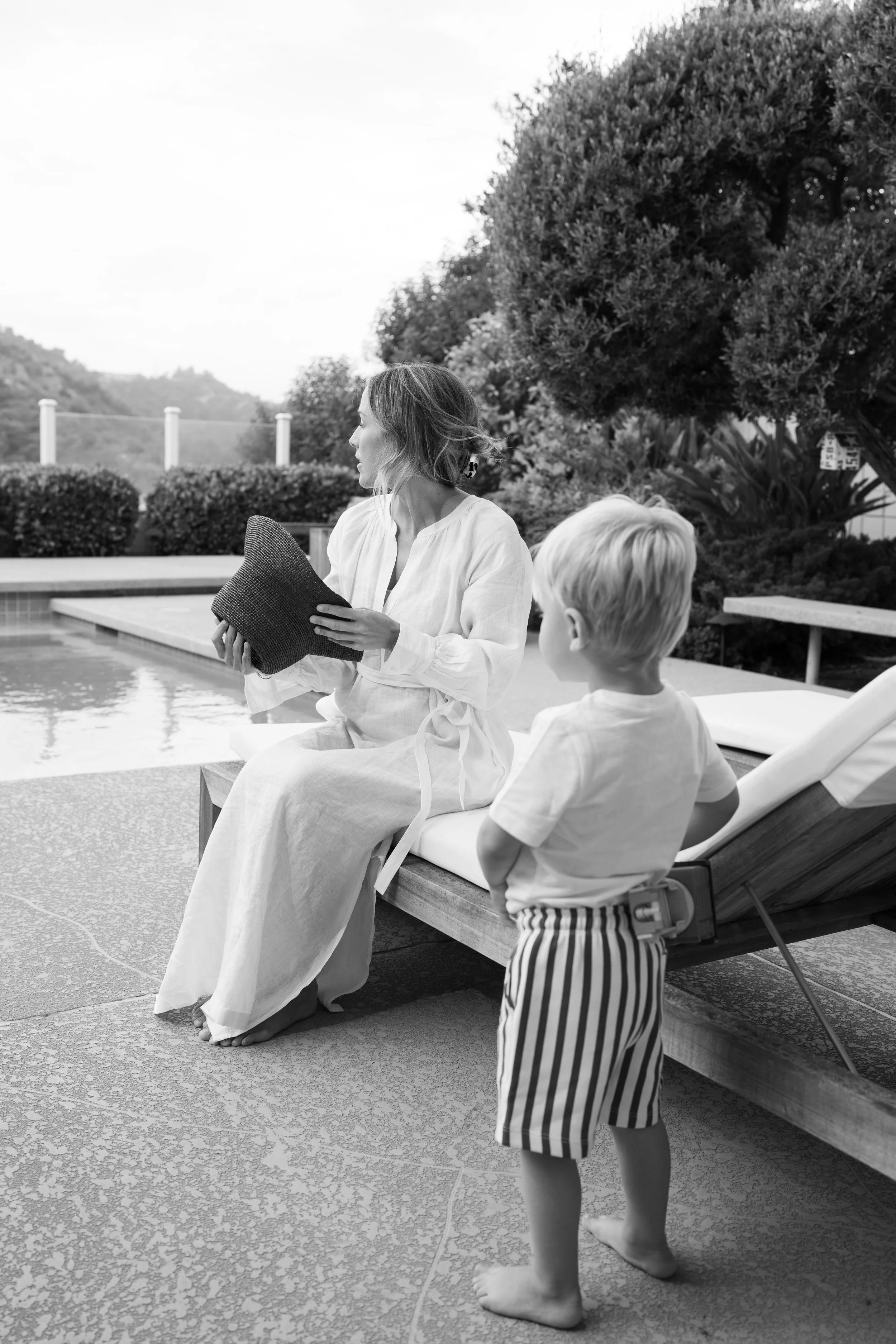 A woman sitting on a poolside lounge chair holding a handbag, with a young boy standing nearby looking at her. The scene is outdoors with trees and mountains in the background.