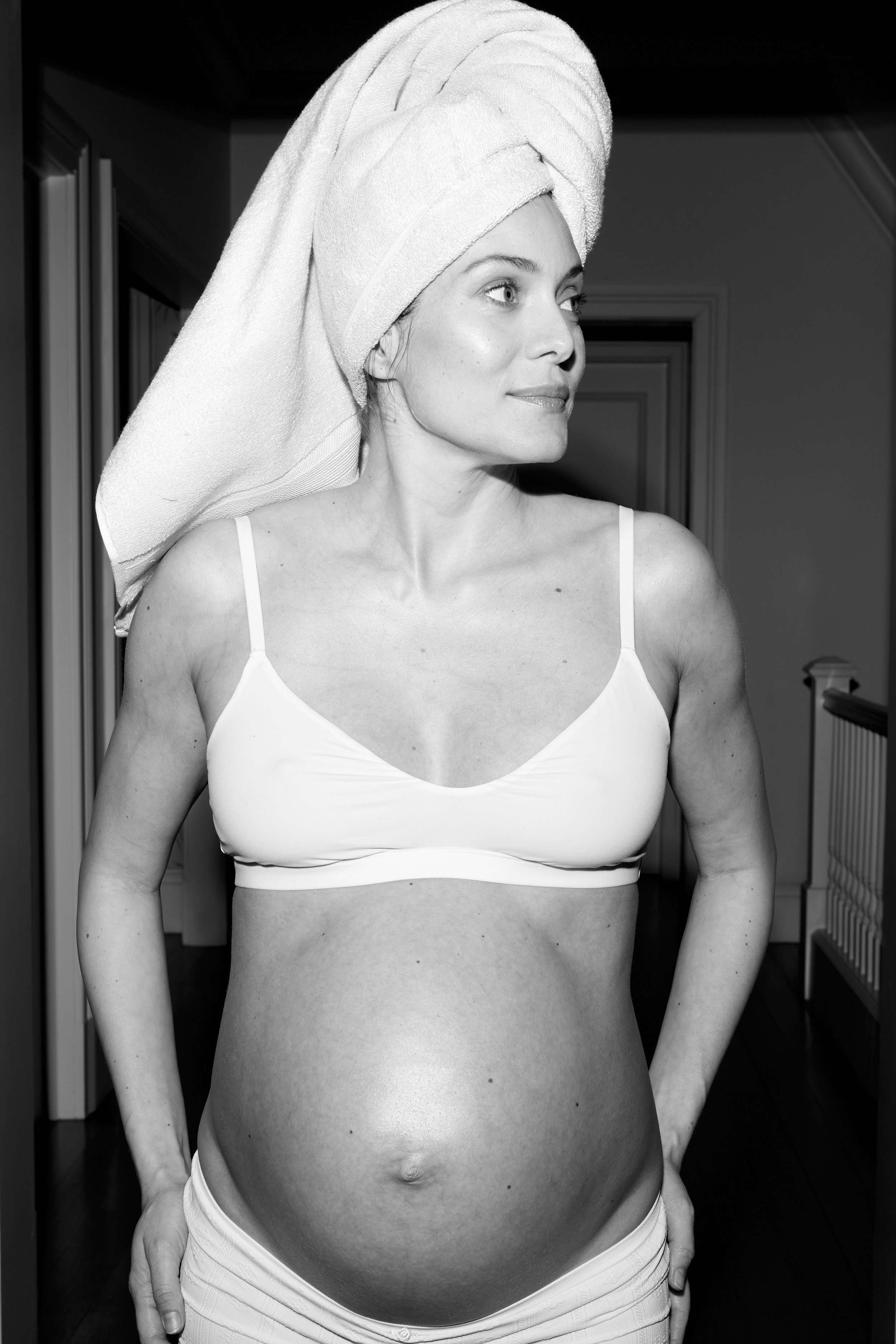 A black and white photo of a pregnant woman with a towel wrapped around her head, wearing a white bra and shorts, standing indoors and looking to the right.