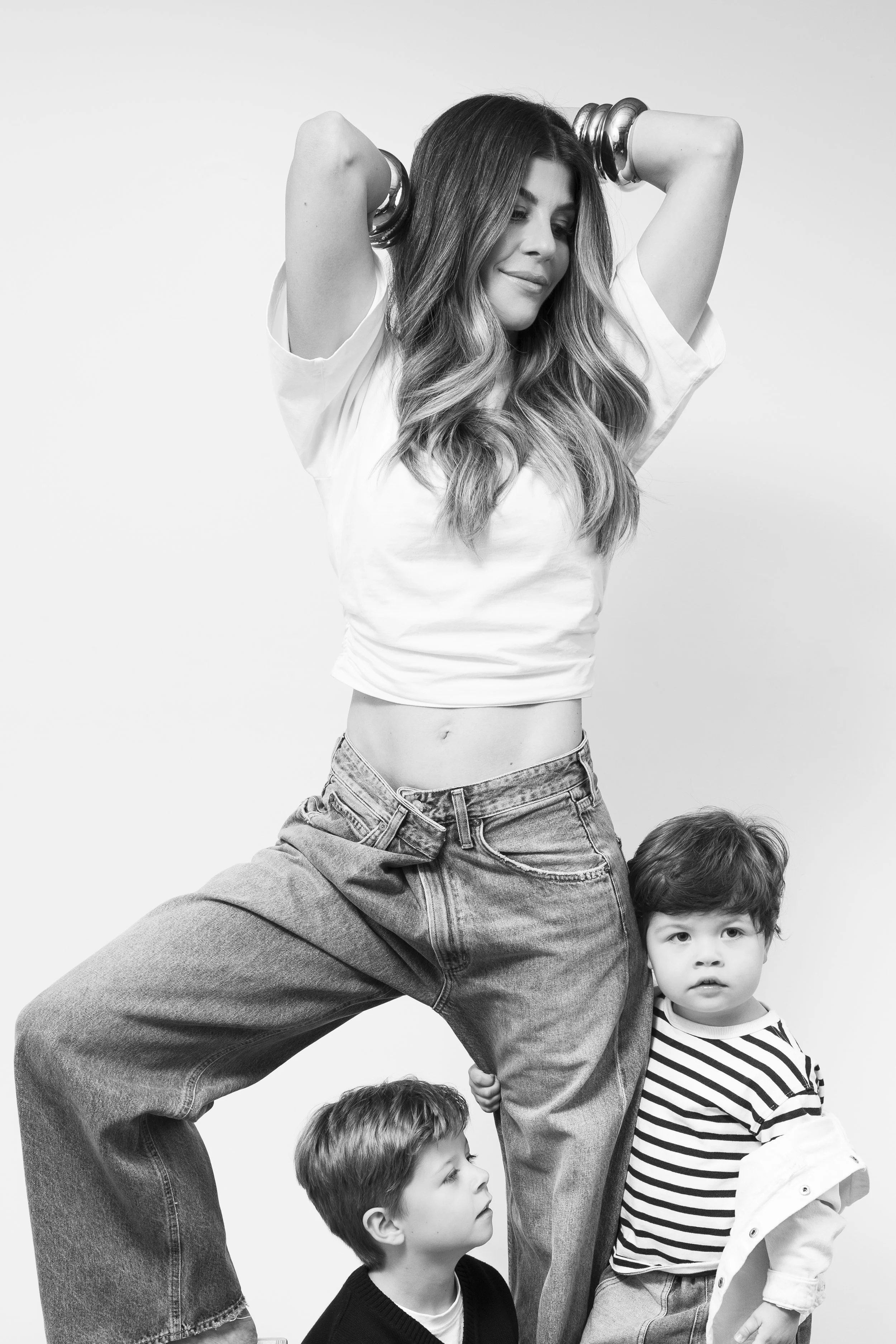 A woman with long hair, wearing casual jeans and a t-shirt, standing with her eyes closed, while two young boys stand next to her, one looking at her and the other looking away. The image is black and white.