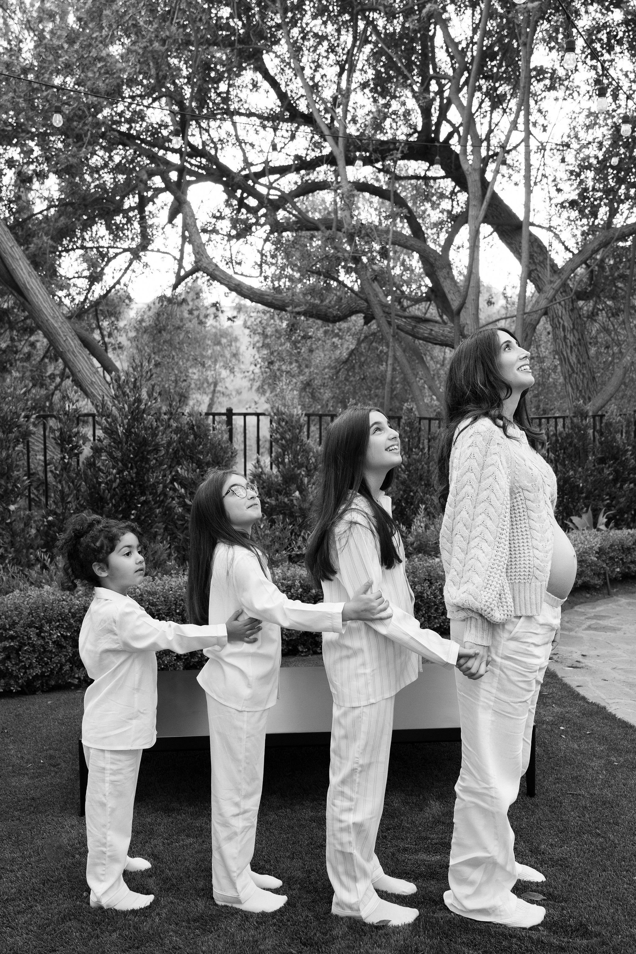 A mother and three daughters stand in a line outdoors, holding hands, looking up and smiling, with a large tree in the background.