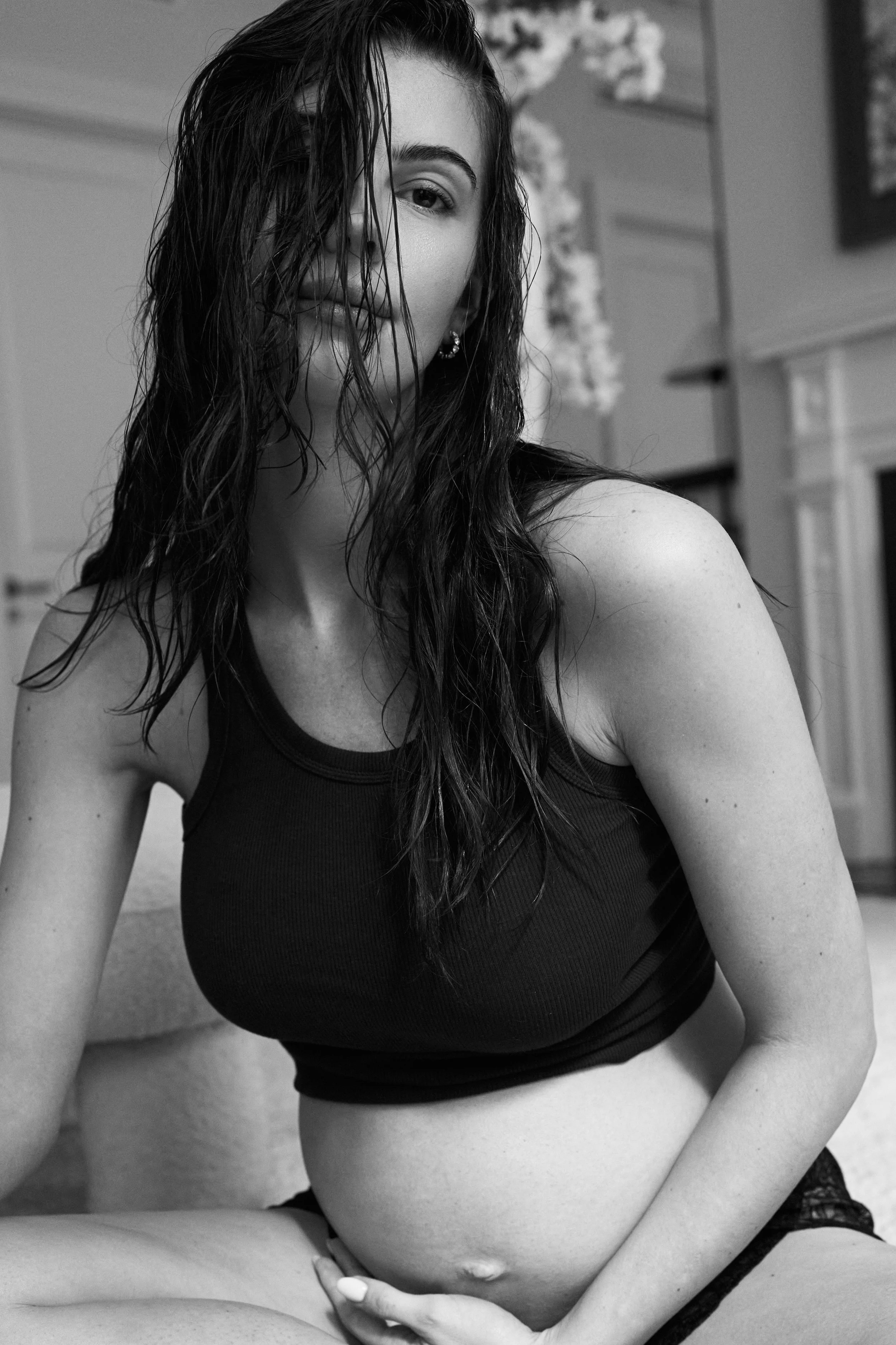 Black and white photo of a pregnant woman with wet hair, wearing a sleeveless black top, sitting on a surface in a home interior.