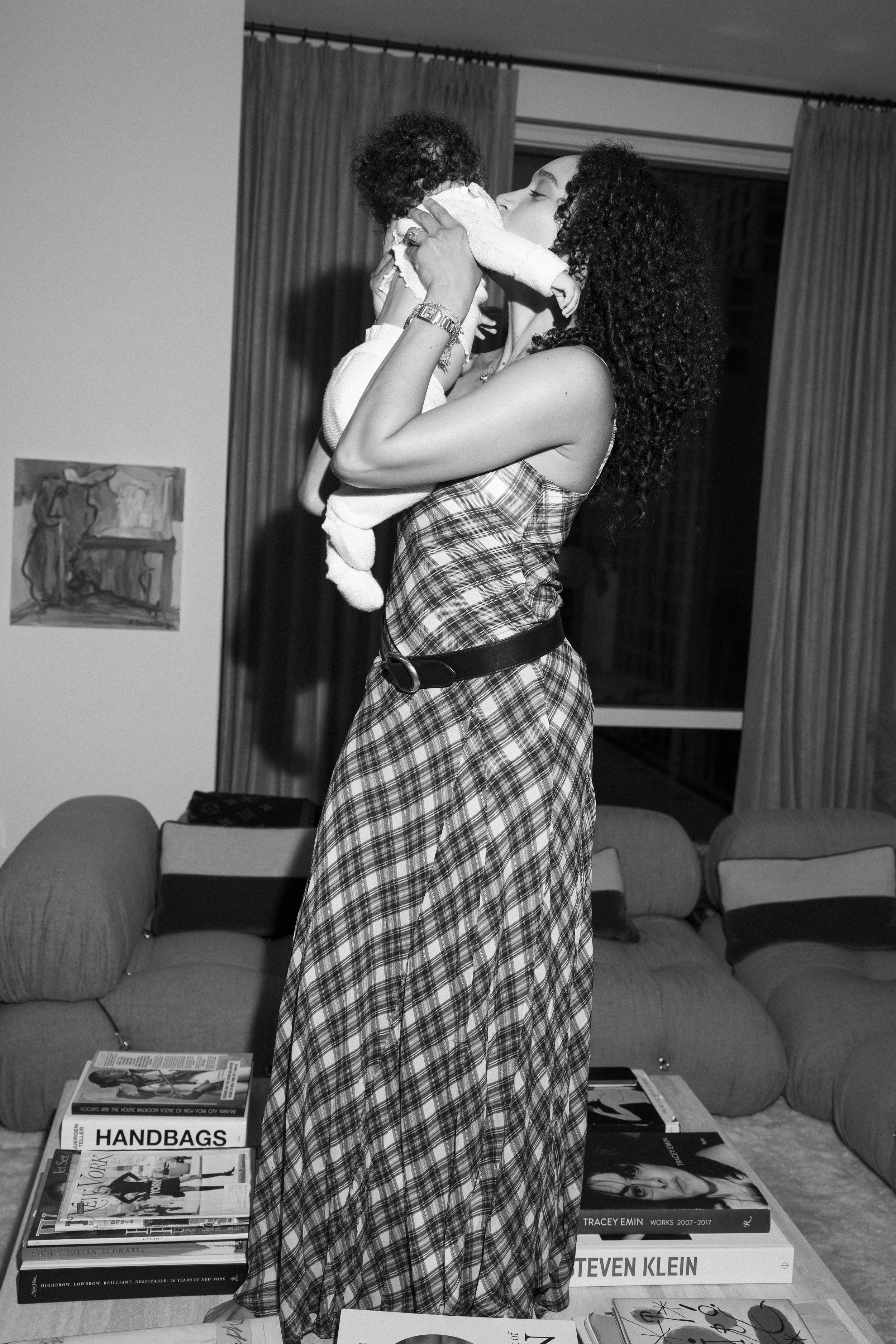 A woman with curly hair holding a baby in a living room with a couch, a table with magazines, and curtains in the background.