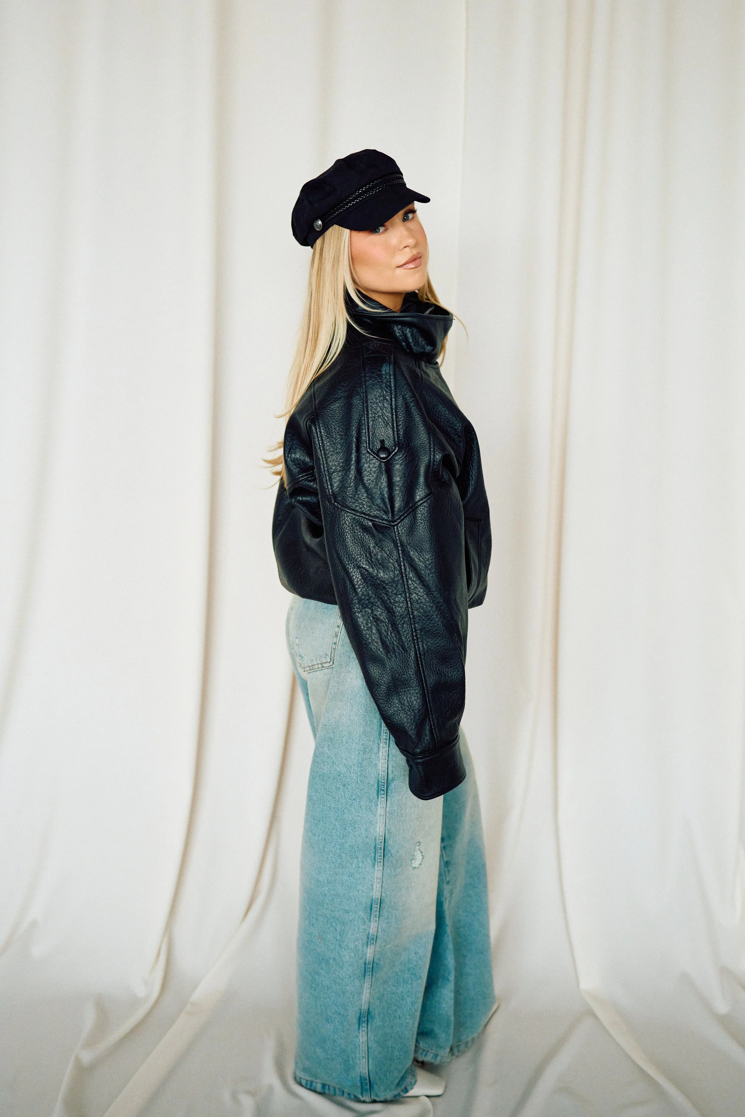 A woman with long blonde hair wearing a black leather jacket, a black cap, light-washed wide-leg jeans, and white shoes, standing against a plain cream-colored background.