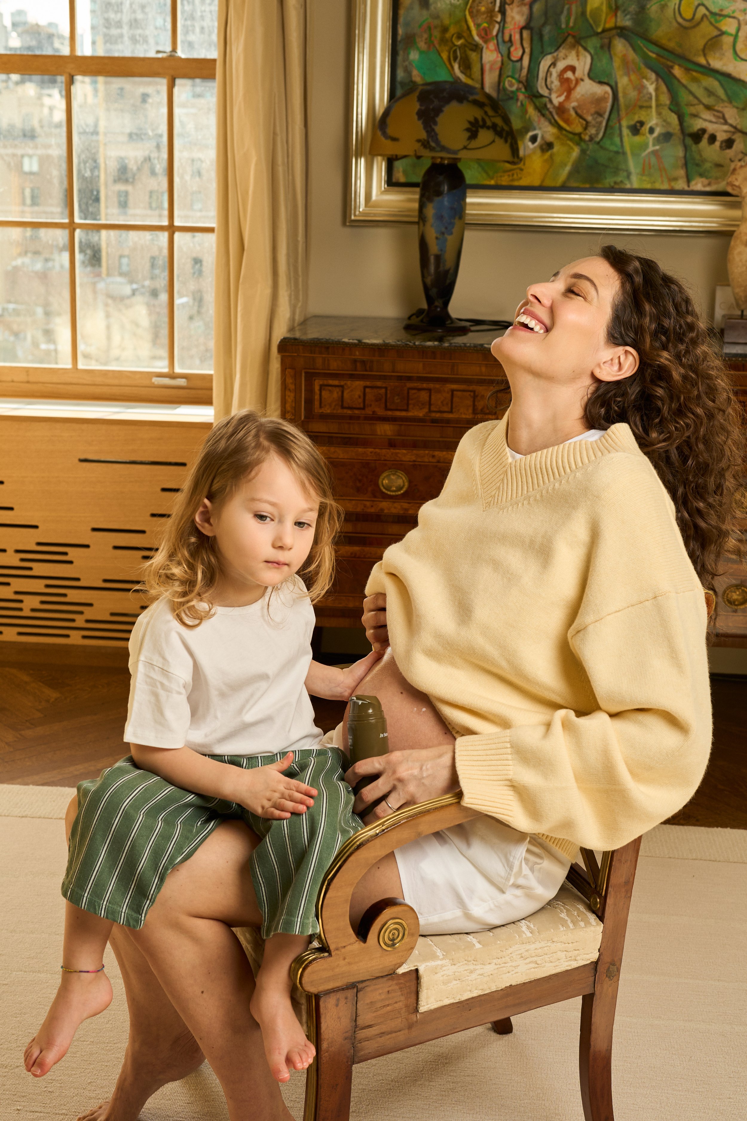 A woman smiling and laughing while sitting on a wooden chair with a young girl on her lap in a warmly lit room with a large window, a vintage wooden dresser, a decorative lamp, and colorful artwork on the wall.