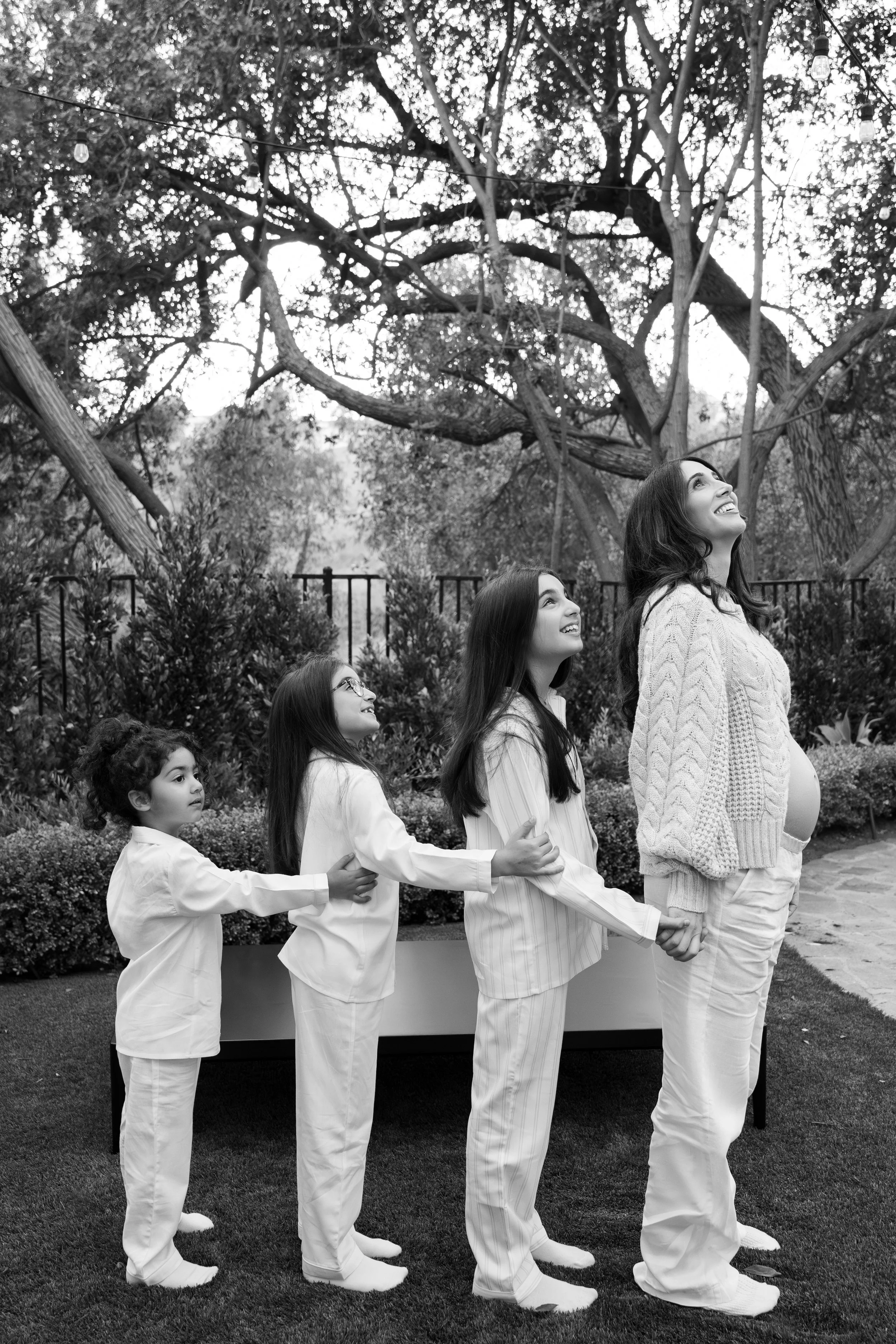A pregnant woman and four young girls, possibly her daughters, standing outdoors in pajamas, holding hands and smiling. The women are looking up, with trees and outdoor lighting in the background.
