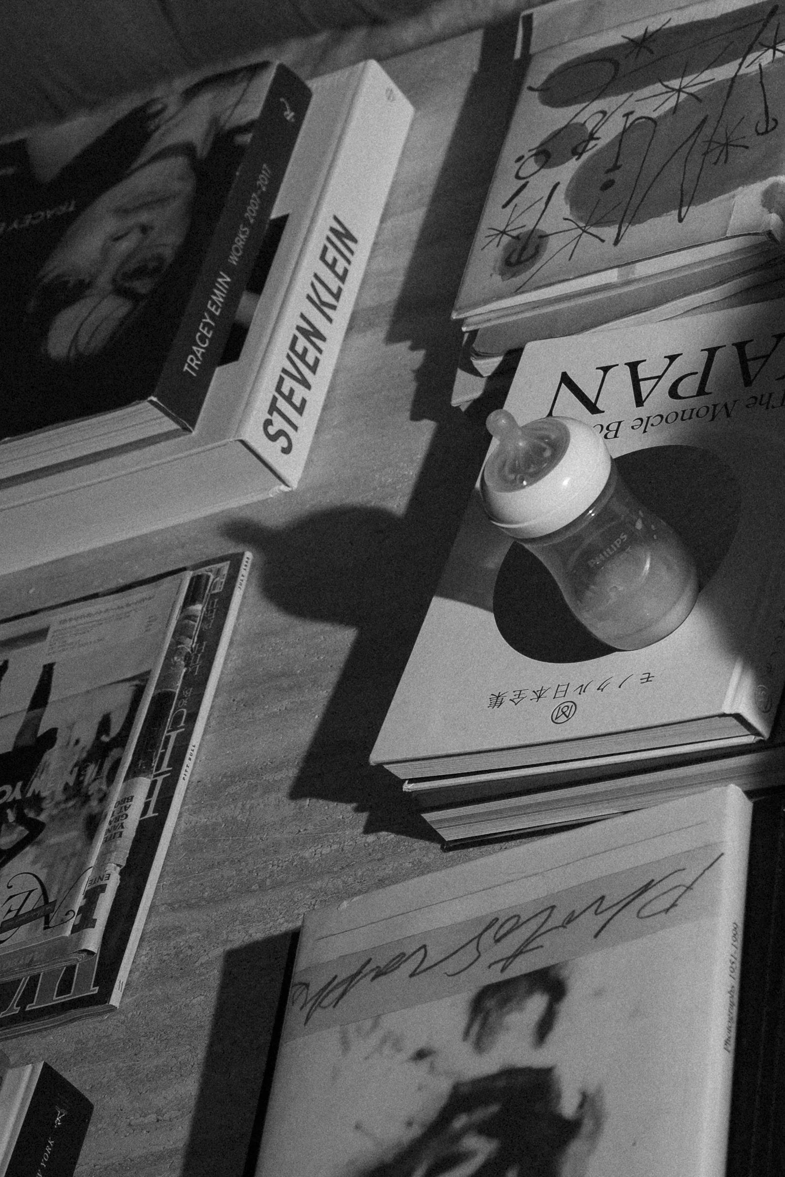 Table with books, a baby bottle, and a magazine seen in grayscale, with shadows cast on the surface.