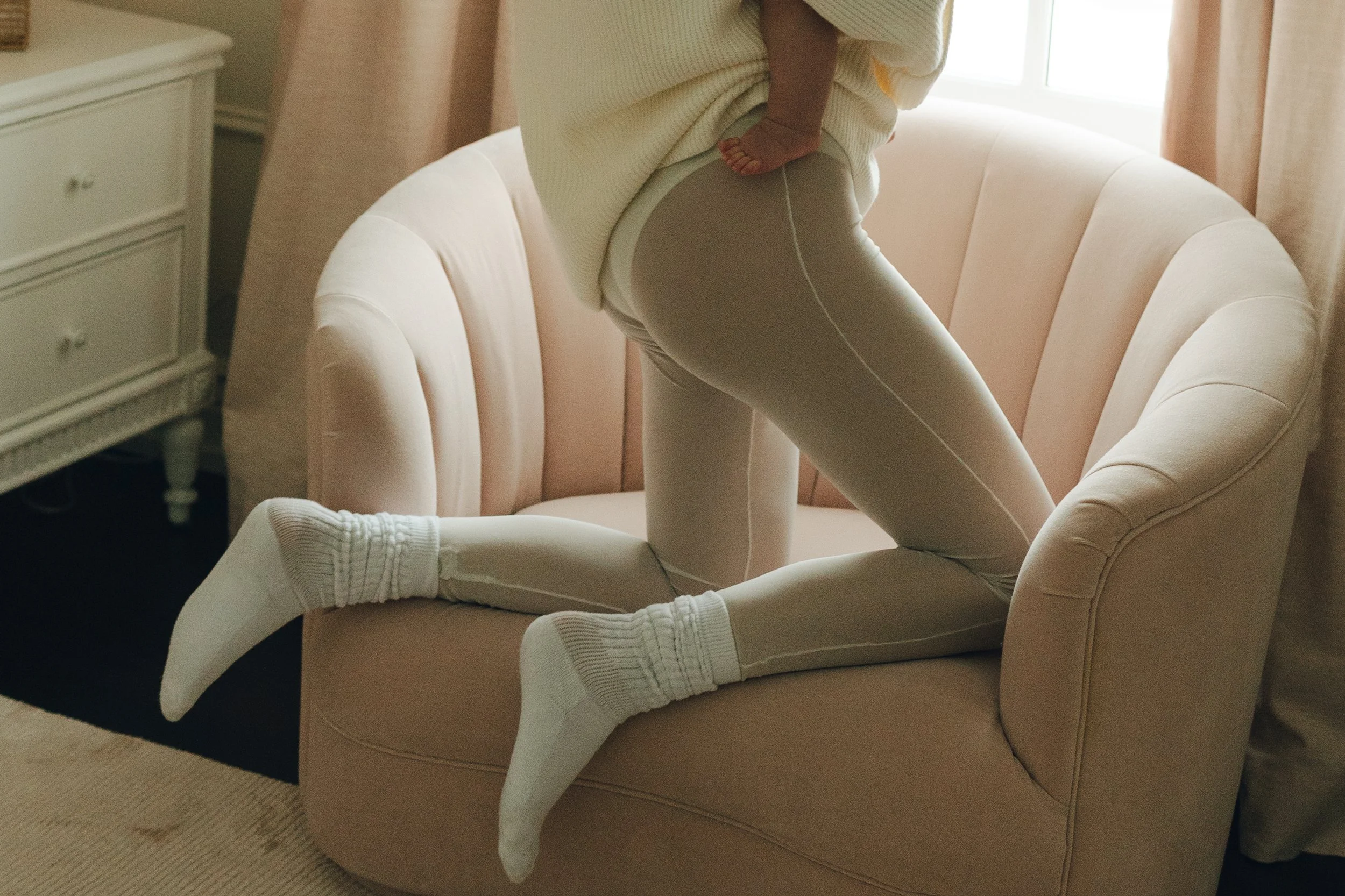 A person kneeling on a pink upholstered chair, wearing beige leggings, a cream-colored oversized sweater, and white socks, with part of a room's interior visible.