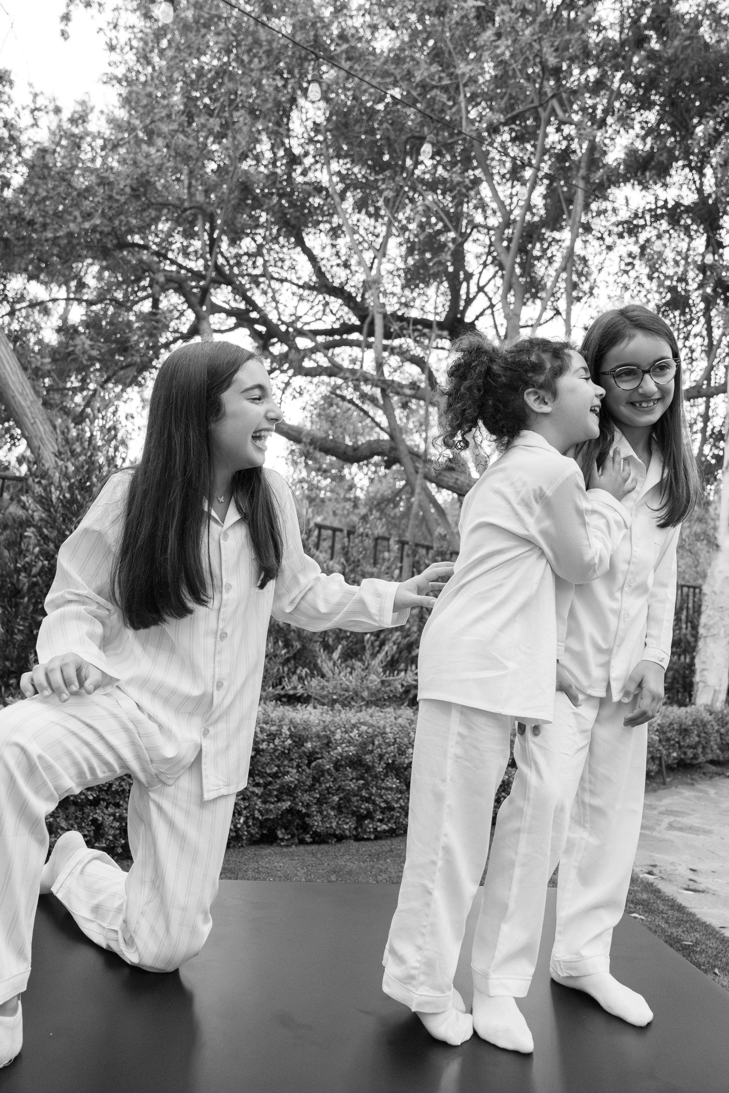 Four young girls with long hair, wearing pajamas, playing and laughing outdoors during daytime, with trees and string lights in the background.