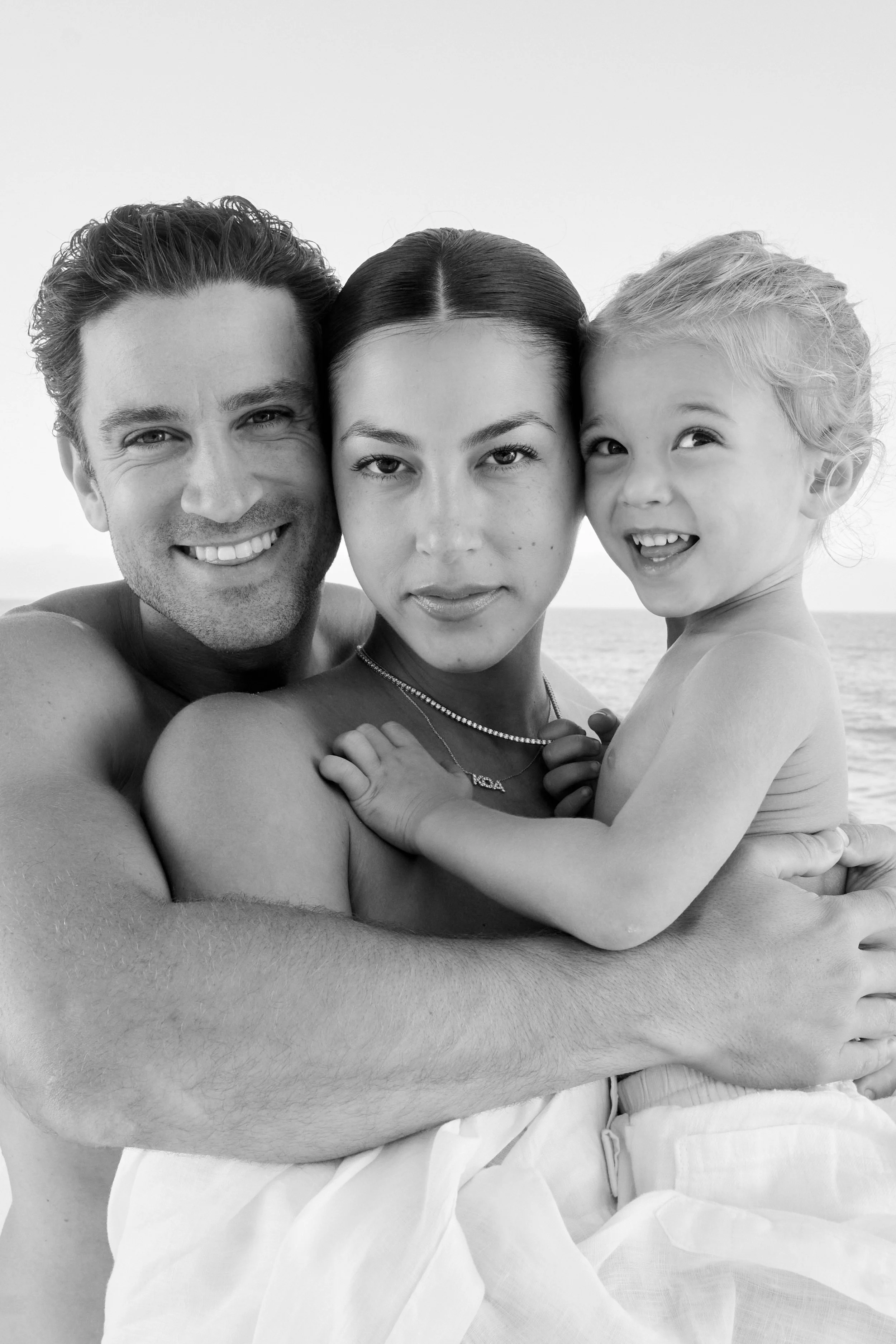 A black and white photo of a smiling man, woman, and a young girl at the beach, embracing each other.