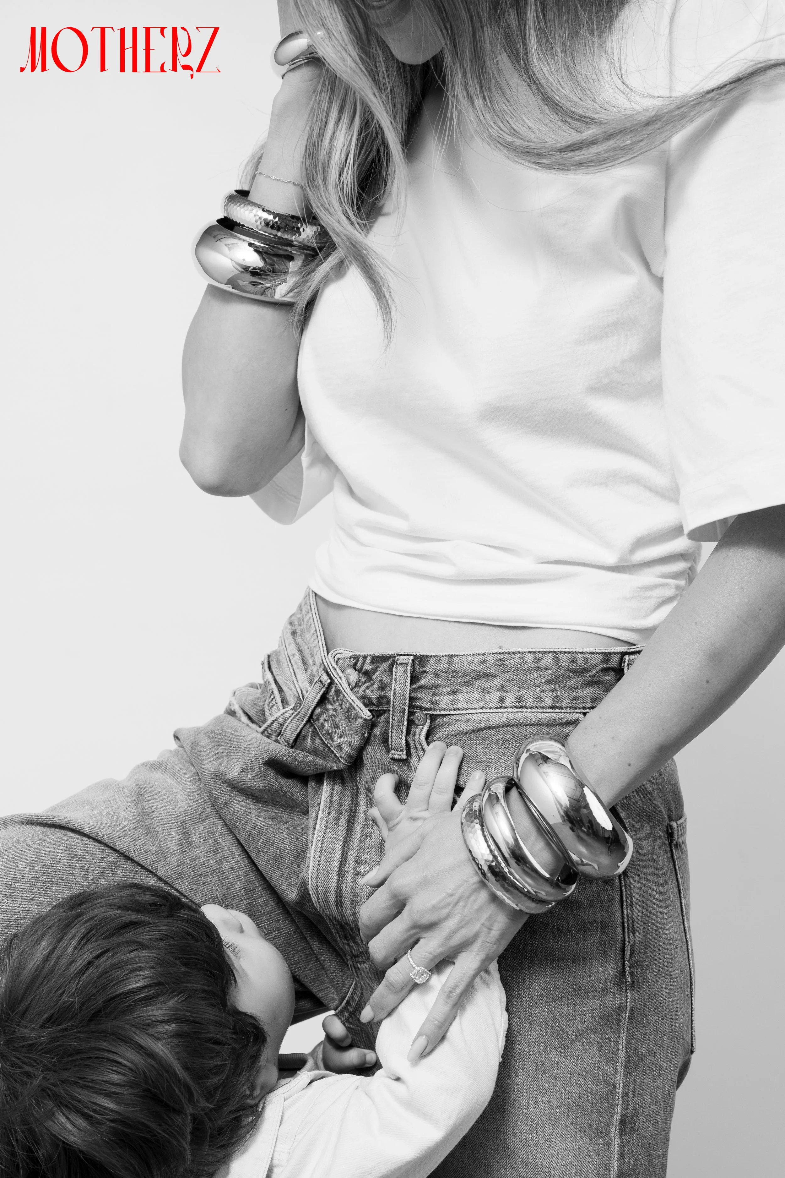 A woman wearing a white t-shirt, jeans, and multiple silver and black bangles on her arm, with a young child holding onto her waist and looking up at her.