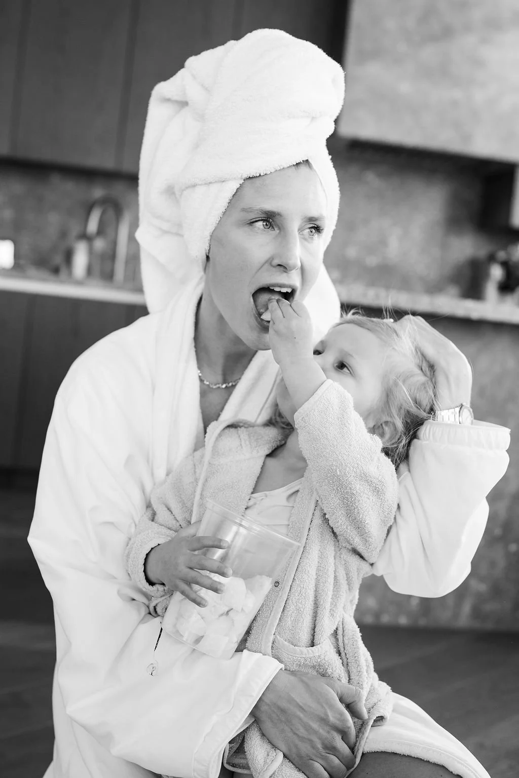 A woman with a towel wrapped around her head playfully biting the fingers of a young girl, who is holding a cup with marshmallows and touching the woman's face. The woman is dressed in a robe and the girl in a fleece jacket, in a kitchen setting.
