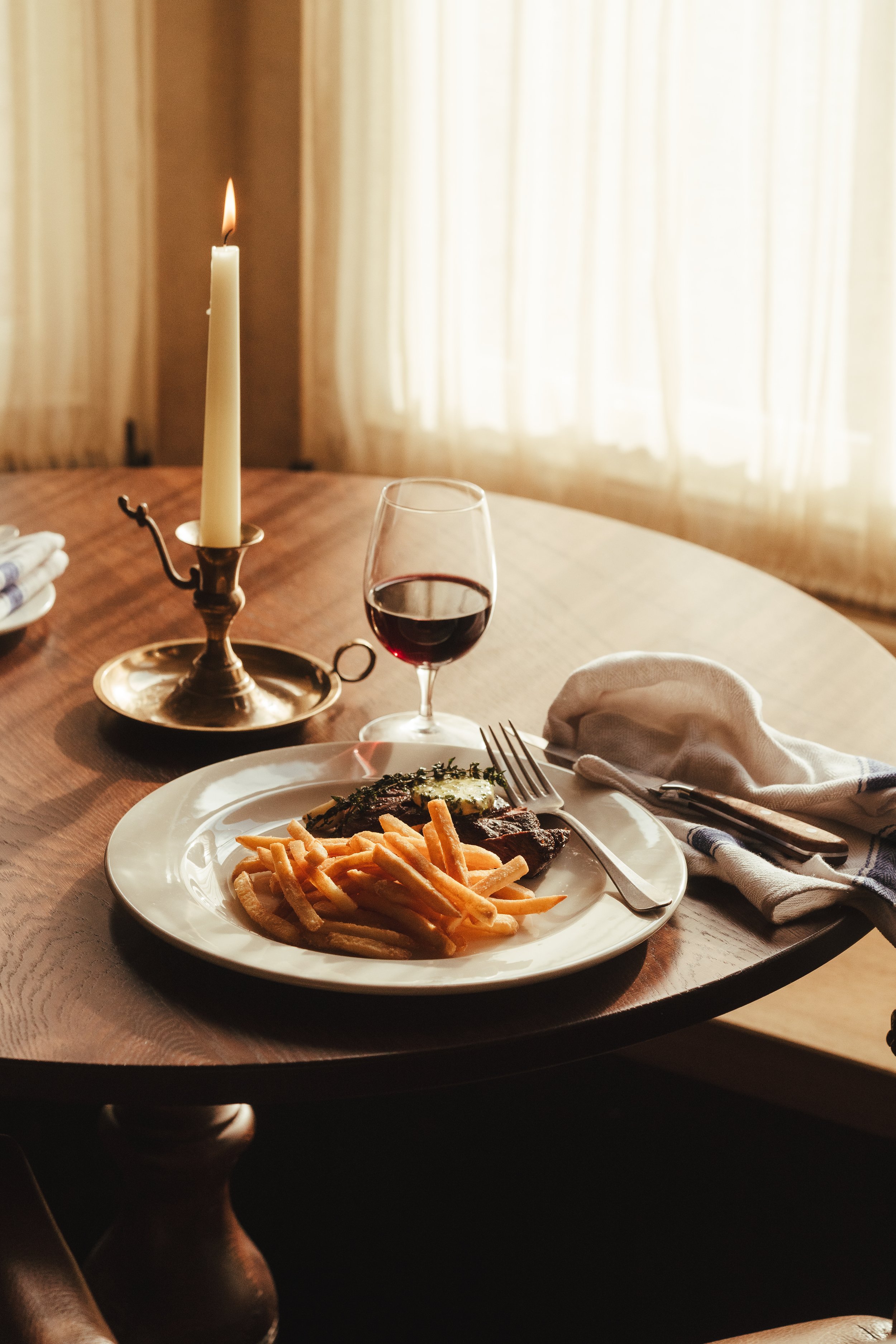 Dinner table set with a lit candle, a glass of red wine, and a plate of steak, fries, and vegetables, with cutlery and a napkin.