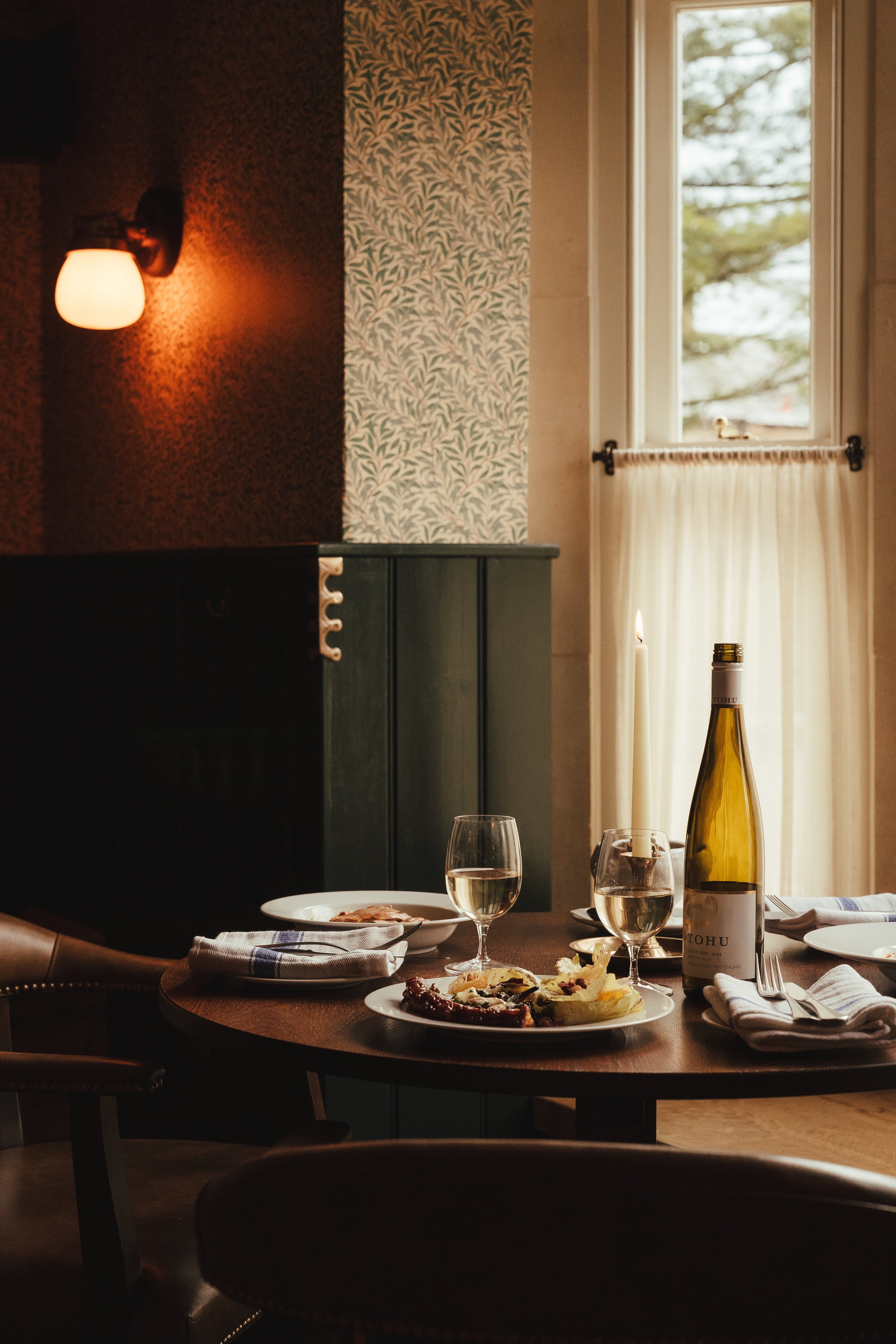 A cozy dining room with a round wooden table set for a meal, including plates with food, glasses of white wine, and a bottle of wine. There are napkins and utensils on the table. A lit candle and a candle holder are on the table. Warm lighting and a 