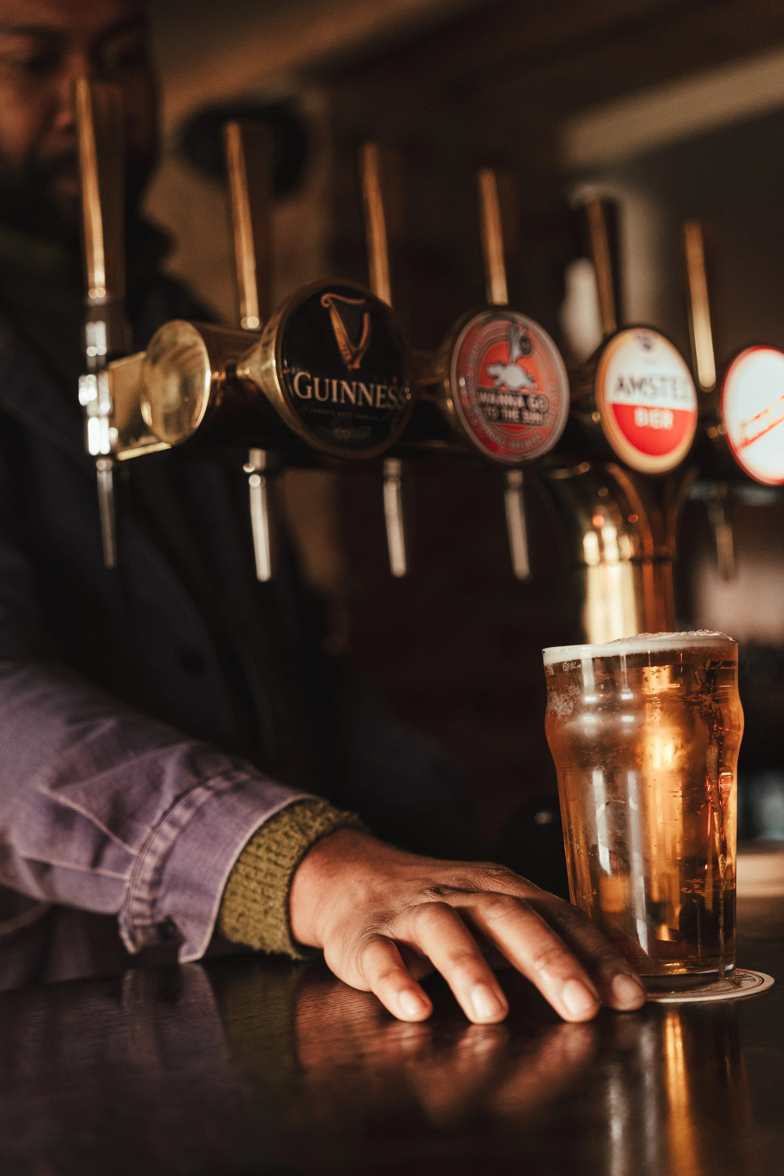 Person reaching for a glass of beer at a bar with beer tap handles, including Guinness, in the background.