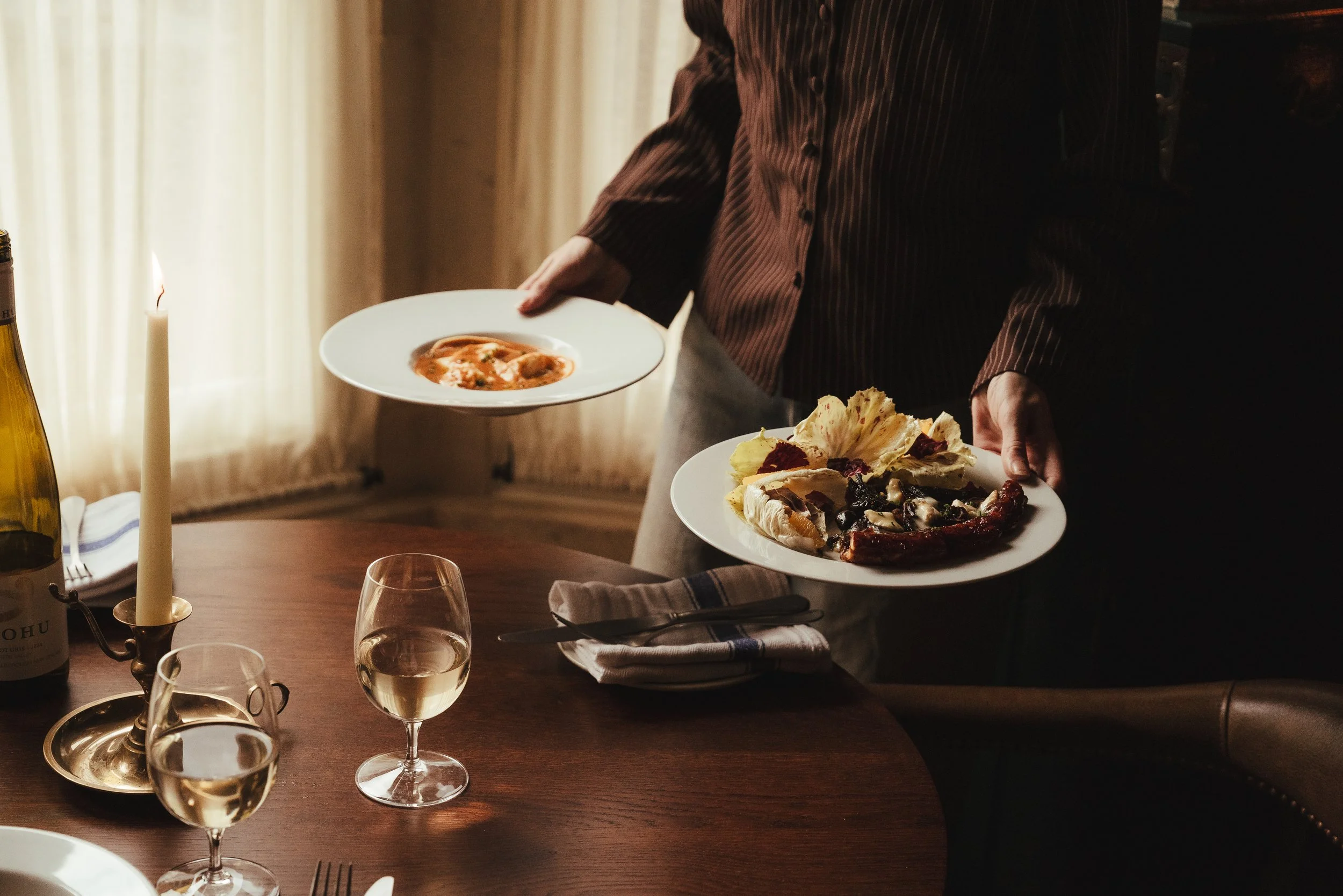 Person serving food at a table with plates of salad and soup, wine glasses, and a candle in a warm, softly lit dining room.