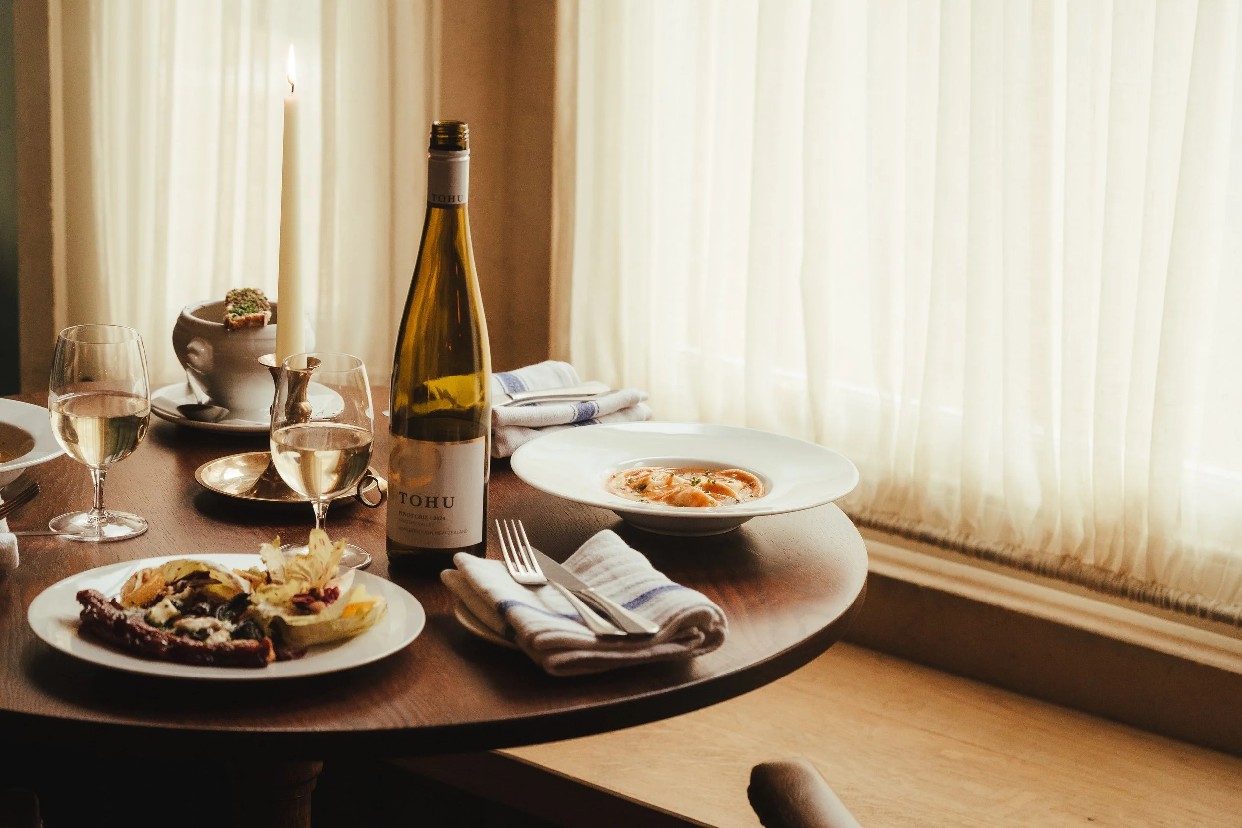A wooden dining table set with white plates of food, a bottle of white wine, glasses of white wine, a candle, silverware, and a napkin in front of a window with sheer curtains.